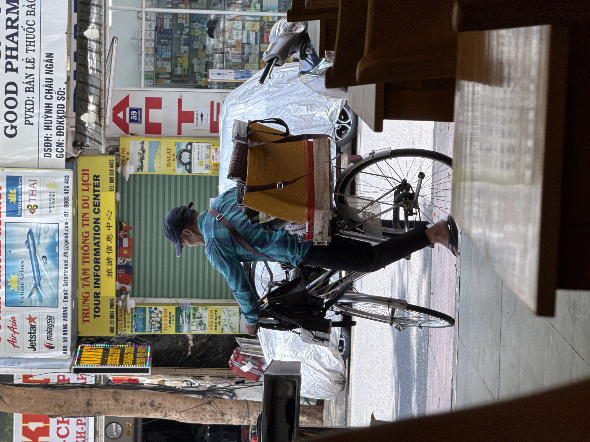 The image depicts a street scene in Nha Trang, Vietnam, specifically in front of businesses. A man, seen from the back, is standing with a bicycle loaded with items. He is wearing a blue and green checkered shirt, dark pants, and dark flip-flops. The bicycle has a large yellow box and a brown bag strapped to the back. In the background, various signs are visible, including advertisements for airlines like "Air Asia," "Jetstar," "Malaysia Airlines," and "Vietnam Airlines." There are also signs for "GOOD PHARM" and "TOUR INFORMATION CENTER" with Vietnamese and Chinese text. A motorcycle is parked to the right, and a car covered with a silver tarp is partially visible behind the bicycle. The environment appears to be a commercial street with buildings lining the road.