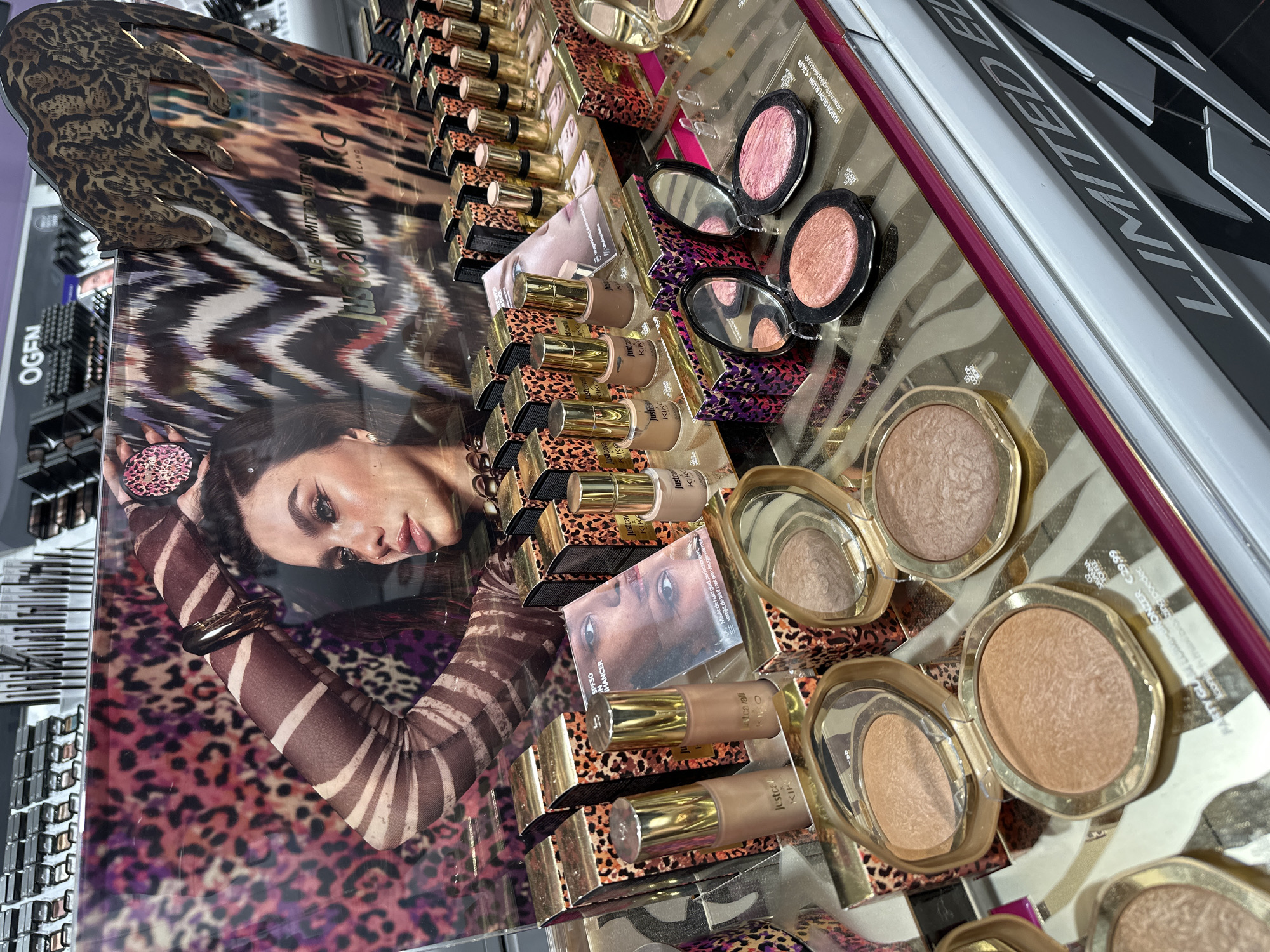 The image shows a display of Kiko Milano makeup products from the Just Cavalli collection, specifically focusing on bronzers, blush, and foundation. The display features promotional images with animal print patterns and a model wearing a similar style. The products are arranged on a glossy surface, possibly a countertop or display case, with additional makeup items visible in the background. The location can be inferred to be in Amsterdam, Netherlands based on the Dutch word "Maakt" visible on the Kiko Milano product packaging.