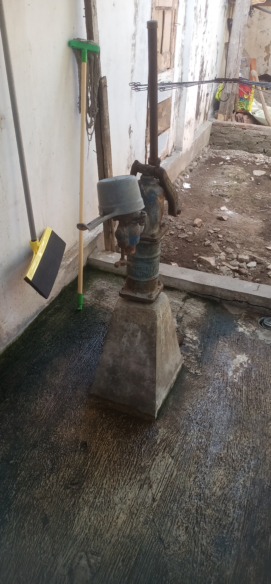 The image depicts an outdoor scene near a wall in Gending, Indonesia. In the foreground, a concrete pad with a textured, wet surface is visible. To the left, leaning against the wall, are a broom with a yellow rectangular head and a wooden handle with a green grip, and a mop with a green head and a wooden handle. A rusty manual water pump stands on a concrete base in the center of the image. The pump has a blue and green body, a spout, and a metal lid covering the top. Behind the pump, a thin, rusty metal pipe extends upwards. The background shows a dilapidated wall with peeling paint and some wooden planks. To the right of the pump, a concrete channel runs along the wall, and beyond that, a dirt and gravel area with scattered rocks and debris is visible. Further back, what appears to be a clothesline with multiple wires and clips is set up. The overall environment suggests an older, possibly rural or less maintained area.
