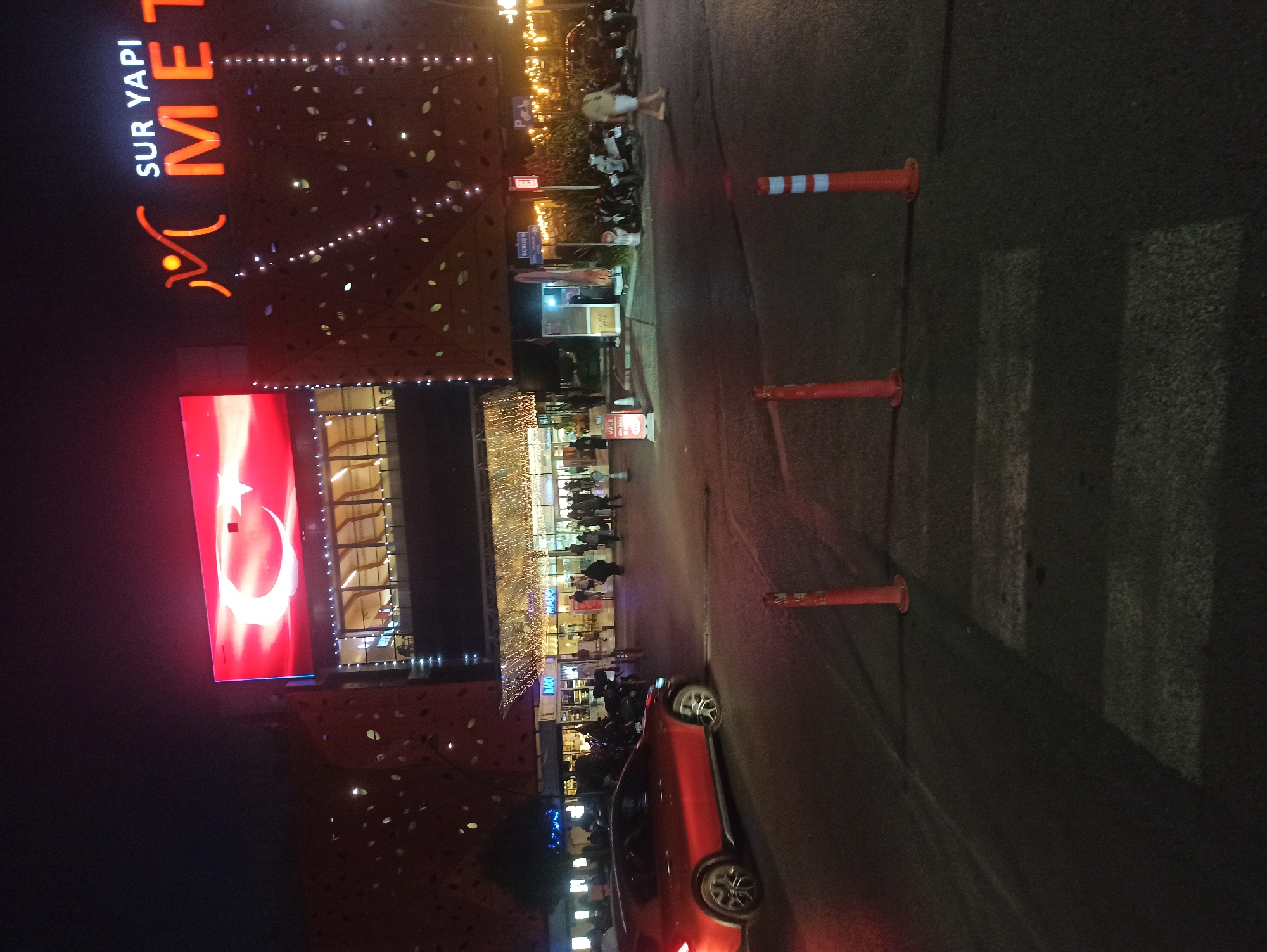 The image captures a night scene at the Metropol Istanbul shopping mall in Ümraniye, Turkey. The mall's exterior is illuminated, with a large LED screen displaying the Turkish flag and the mall's name, "SUR YAPI METROPOL," in prominent signage. The building's facade is decorated with intricate, perforated patterns and strings of lights.
In the foreground, a red car is parked on the street, with its headlights on. Several people are visible walking in front of the mall entrance, some appearing to be entering or exiting. Motorcycles are also parked near the entrance. Traffic cones and a pedestrian crossing are present on the road. The overall atmosphere suggests a busy, well-lit urban environment at night.