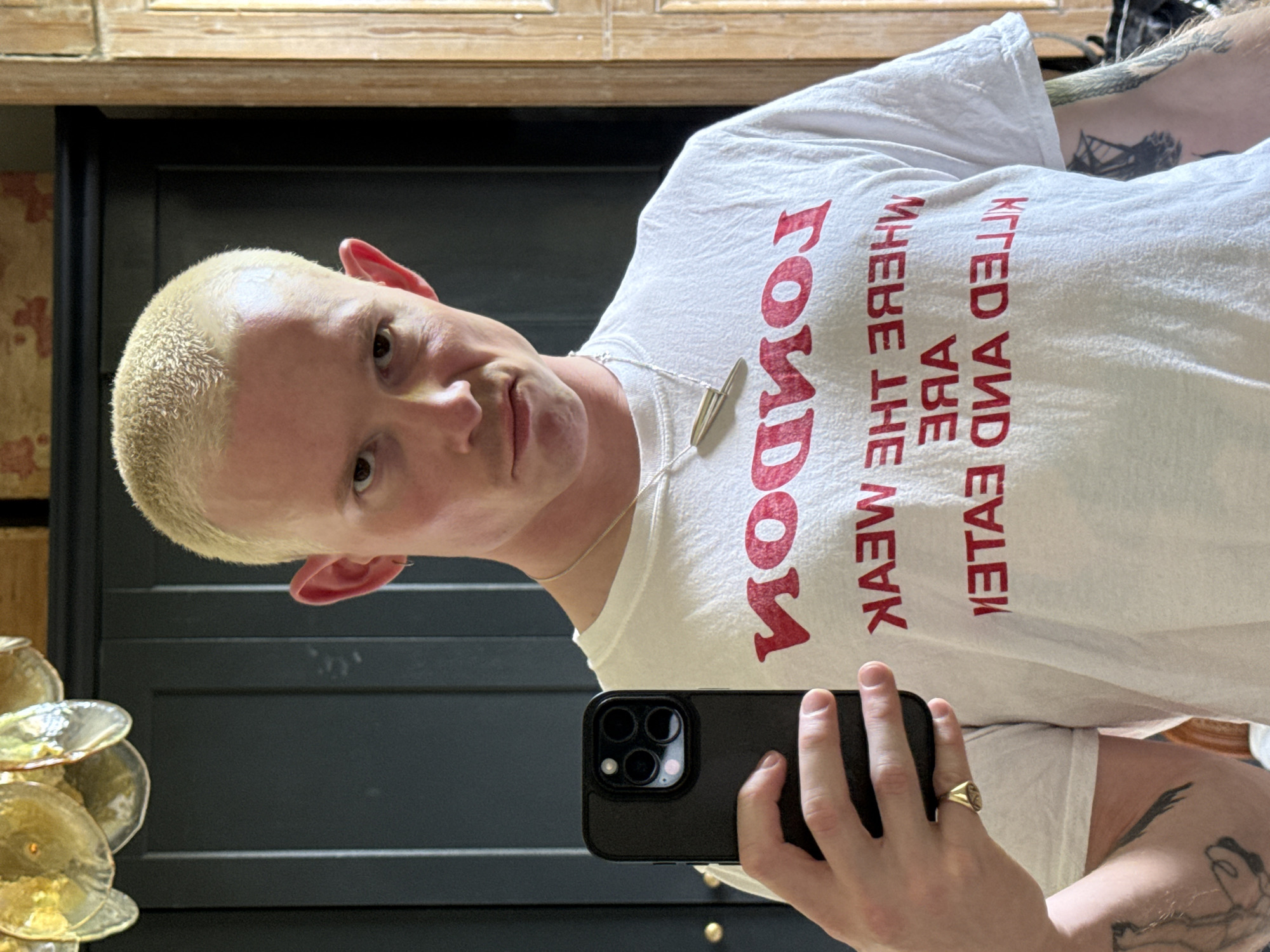 A person with light, buzzed hair and fair skin takes a mirror selfie indoors. The individual is wearing a white t-shirt with red, mirrored text that reads "LONDON WHERE THE WEAK KILLED AND EATEN." A silver-colored necklace with a geometric pendant hangs around their neck. They are holding a black smartphone, likely an iPhone Pro model with a triple camera system, in their left hand, which also displays a gold-colored signet ring on the ring finger. Tattoos are visible on the right arm and shoulder area.
The person has a neutral expression, looking directly into the phone/mirror. The background features dark paneling or furniture, possibly a fireplace surround, with a lighter wooden surface above it. To the lower left, part of a decorative art piece or light fixture made of multiple, layered circular or leaf-shaped elements is visible. The lighting appears bright and natural, suggesting it is daytime. The image is taken at a slight angle and is rotated ninety degrees counter-clockwise. While the shirt mentions "LONDON," there are no other explicit visual cues in the image to confirm the specific location within the city.
