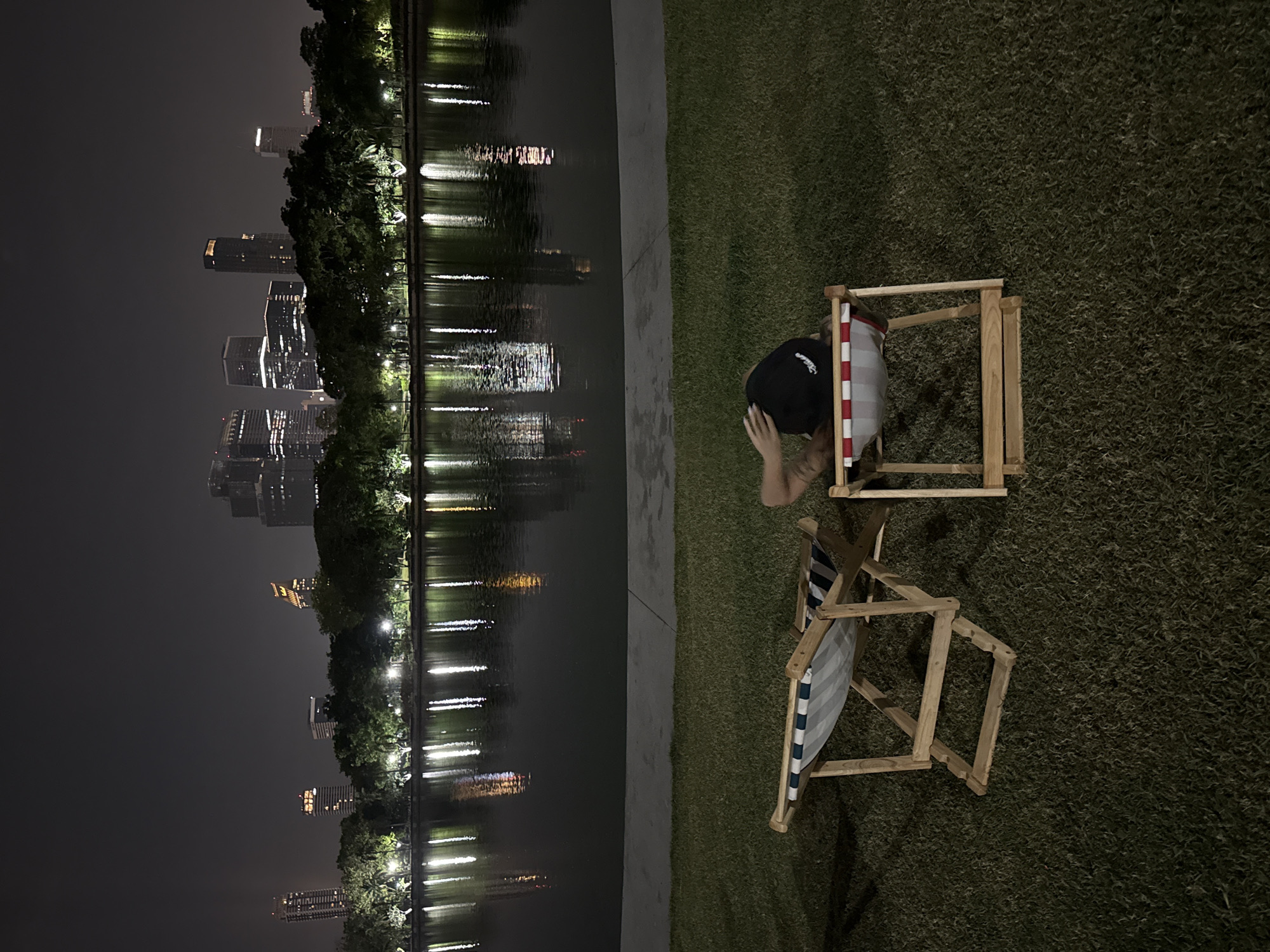 The image depicts a nighttime scene along a body of water, likely a lake or river, in Bangkok, Thailand. The cityscape is visible in the background, with illuminated skyscrapers reflecting in the water, suggesting a calm surface.
Two empty deck chairs made of wood and fabric with striped patterns (one with red and white stripes, another with blue and white stripes) are positioned on a patch of grass, suggesting a place for relaxation or leisure. To the right of the chairs is a person wearing a black cap sitting on a deckchair. Their hands are placed on their head. A concrete walkway separates the grass from the water's edge. The overall lighting is dim, with artificial lights from the city and possibly streetlights providing the illumination. The image appears to be taken from an elevated perspective, looking down towards the chairs, water, and cityscape.