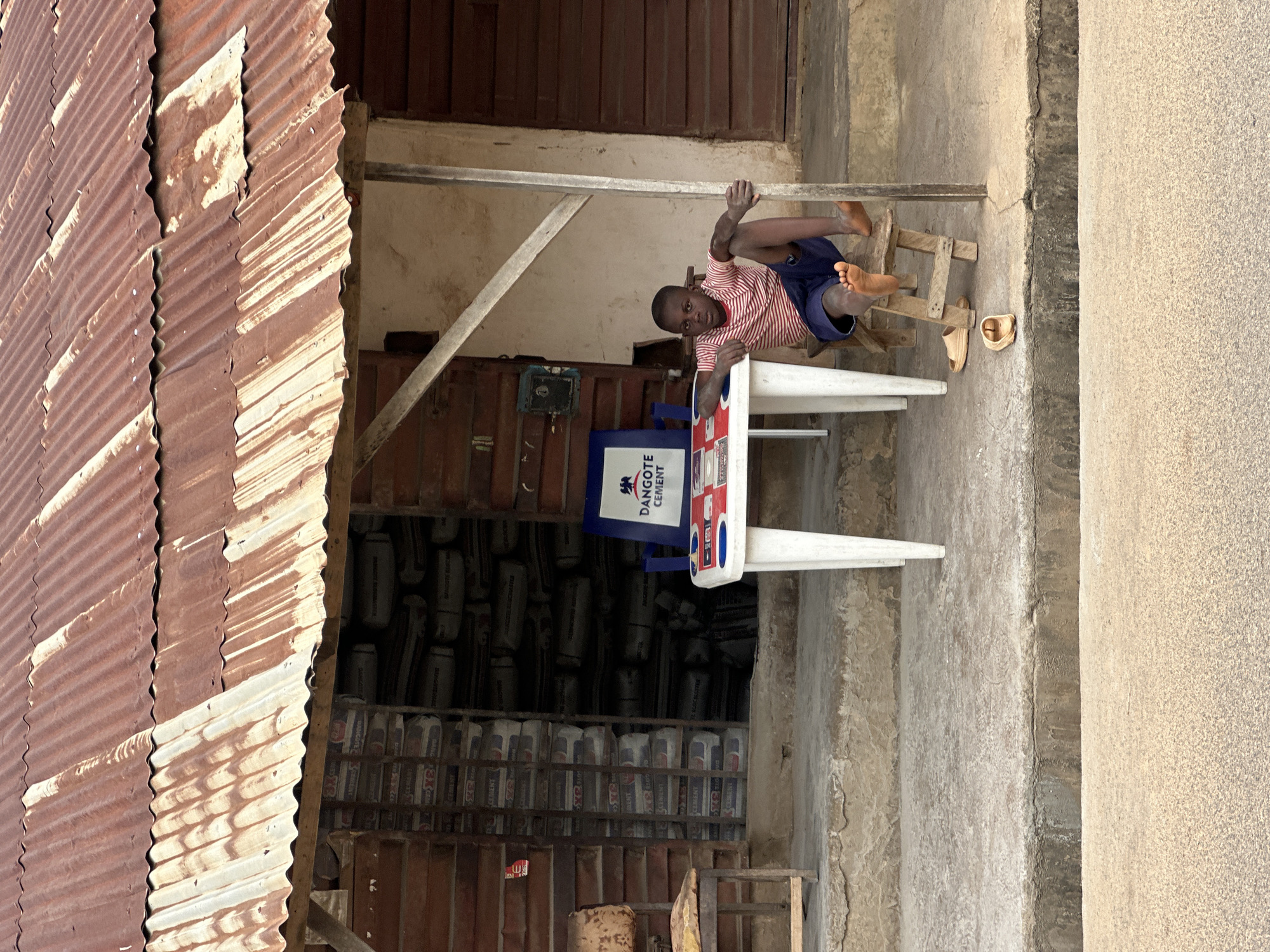The image depicts a young boy in Akwanga, Nigeria, lounging on a small wooden stool. He is wearing a red and white striped t-shirt and blue shorts. The boy is positioned near a white and blue table displaying a sign for "Dangote Cement." Behind him, shelves are stocked with bags of cement. The setting appears to be an outdoor shop or storage area, with a corrugated metal roof on the left and a concrete floor. Wooden structures are visible throughout the scene. The lighting suggests it is daytime, and the atmosphere seems relaxed, with the boy appearing comfortable and perhaps bored. His casual posture and the presence of sandals on the ground indicate a warm, informal environment.