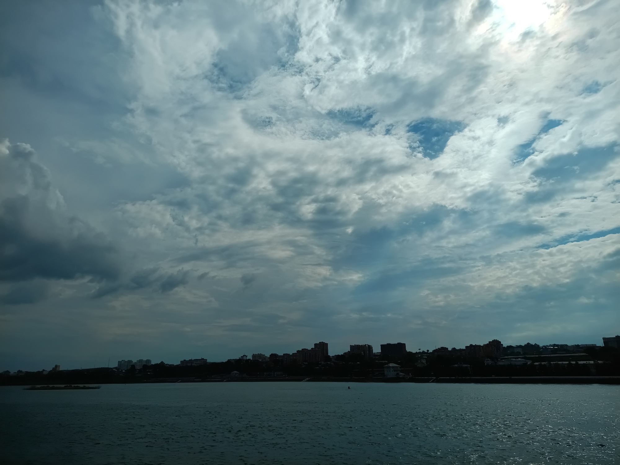 The image depicts a wide, choppy body of water in the foreground, likely a river or lake, reflecting the sky. In the distance, a silhouetted city skyline is visible against a dramatic sky filled with large, textured clouds. The clouds are a mix of dark gray and bright white, with patches of blue sky peeking through, suggesting a dynamic weather pattern. The sun is partially obscured by the clouds, creating a bright glow in the upper right portion of the image. The city appears to be situated on a gentle slope rising from the water's edge, with numerous buildings of varying heights. The overall impression is one of a vast natural landscape meeting an urban environment under a dramatic sky. This scene is consistent with views of the Angara River in Irkutsk, Russia, which flows through the city and is bordered by its urban development and dramatic cloud formations, especially during transitional weather periods. There are no people or discernible actions visible in the image.