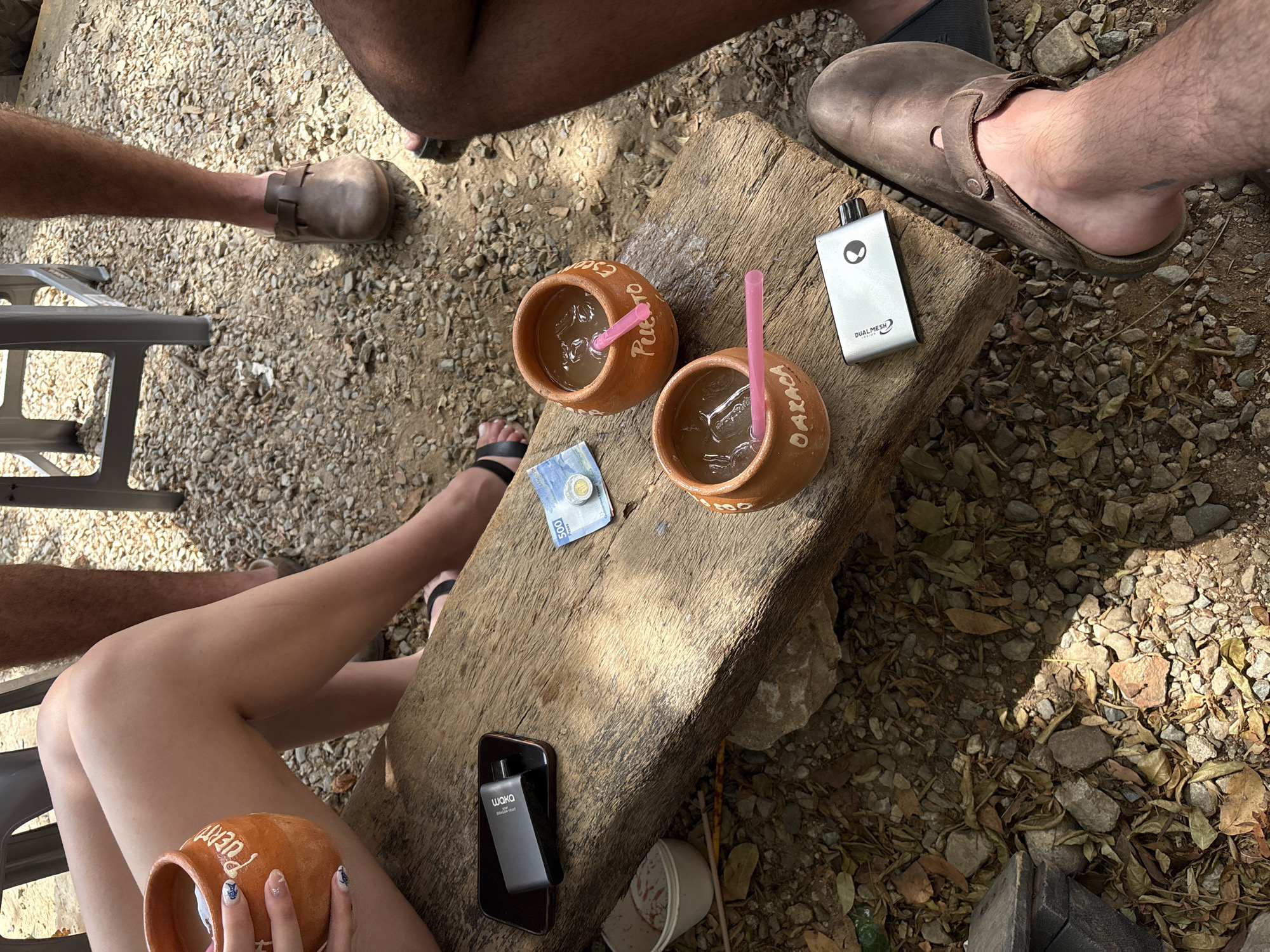 The image depicts an outdoor social gathering, likely a meal or drinks, in Brisas de Zicatela, Mexico. Four people are partially visible. One person holds a clay cup inscribed "Puerto," and another clay cup is visible with "Oaxaca." A 500 Mexican peso bill lies on the table next to the two cups. There is an e-cigarette, a phone and a receipt. The environment includes a gravel ground with leaves.