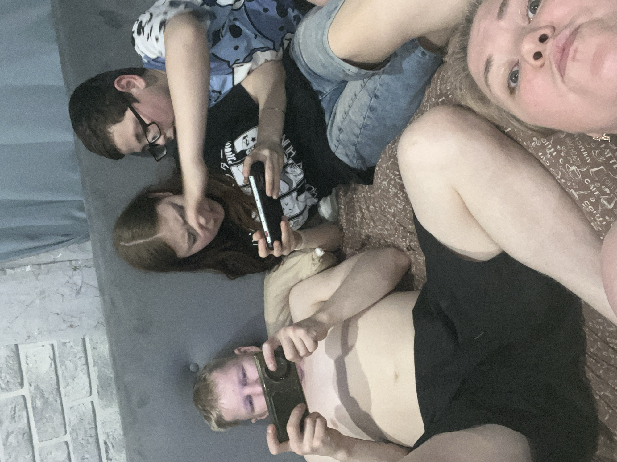 Four individuals are lying down, oriented in different directions. Three are holding and looking at cell phones. A male with glasses is wearing a blue patterned shirt and jeans. A female with long brown hair is wearing a black shirt. A male with blond hair is shirtless and wearing black shorts. A blond female is visible in the top right corner of the image. The background includes a grey surface resembling a sofa and a brown patterned surface possibly a bed. The scene appears to be indoors. There is no verifiable information to suggest that this photo was taken in Vesyoloye, Russia.