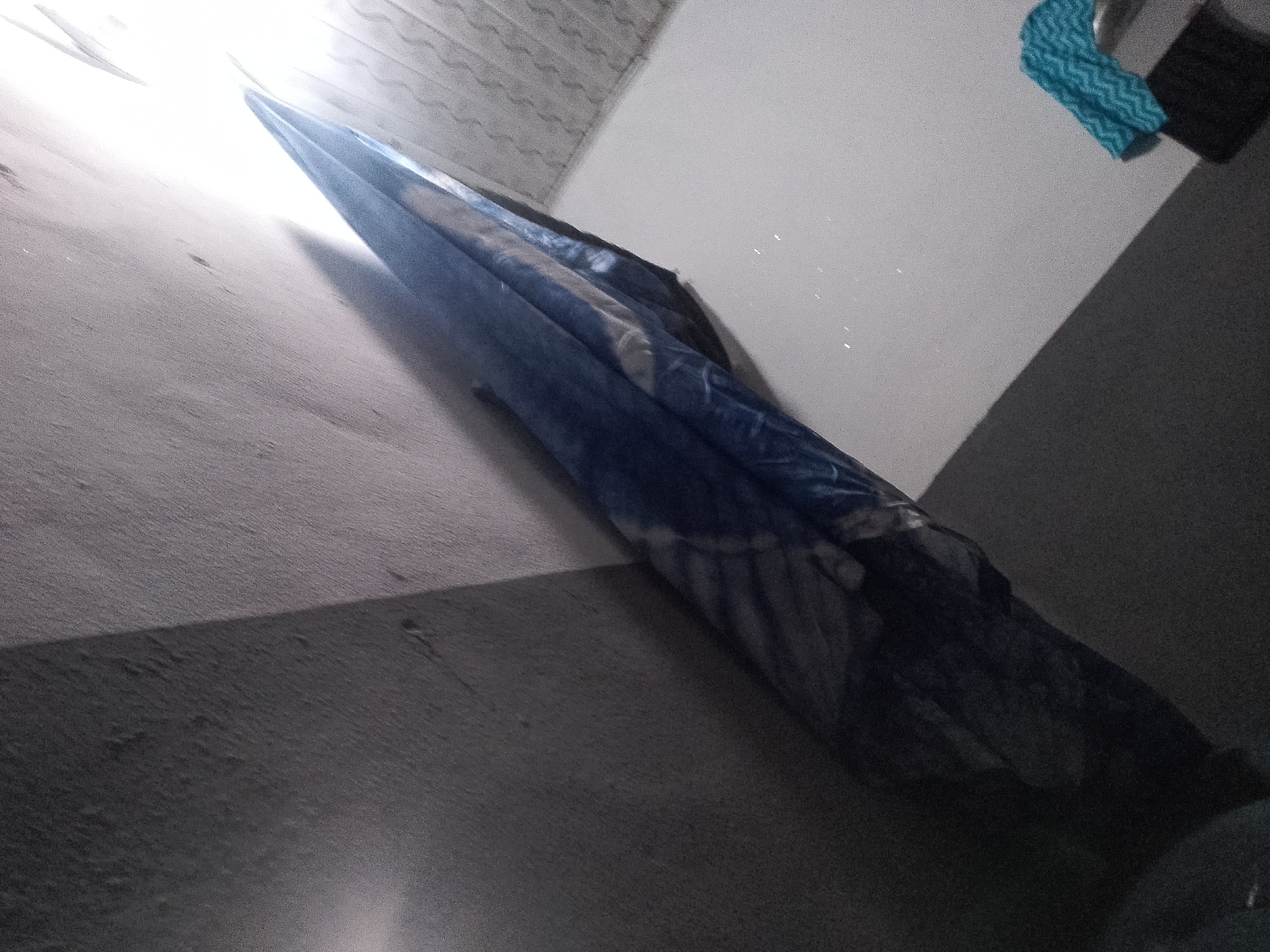 An indoor, low-angle shot shows a blue and white striped blanket hanging from a wall in what appears to be a closet or narrow room. The blanket is draped over an unseen object, creating a long, pointed shape that extends diagonally across the frame. A bright light source is visible at the top left of the image, casting a shadow down the wall. In the upper right corner, a blue textured towel and a black object are visible. The overall lighting is dim, with the exception of the light source at the top.