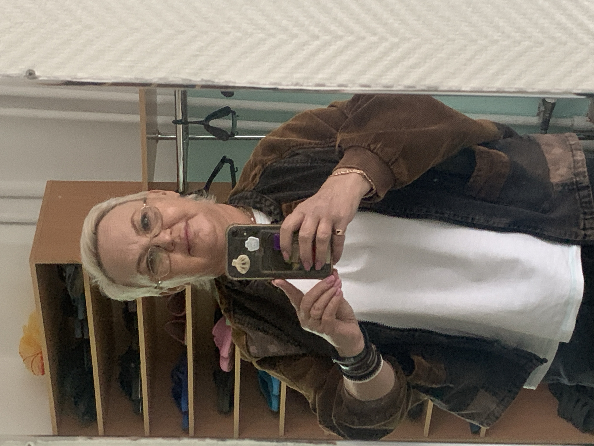 A woman with short, light blonde hair and eyeglasses is taking a selfie in a mirror. She is wearing a white t-shirt and a brown corduroy jacket with darker patches. She holds a smartphone in her right hand, the phone's case adorned with stickers. The mirror reflects a setting that appears to be indoors, possibly a public space or hallway with wooden cubbies/shelving units. Behind the woman, visible through the mirror's reflection, are clothing items and other miscellaneous objects stored in the shelves. Above the shelving units is a metal rod and clothing hangers. There is no verifiable information to confirm this image was taken in Chaikovsky, Russia.