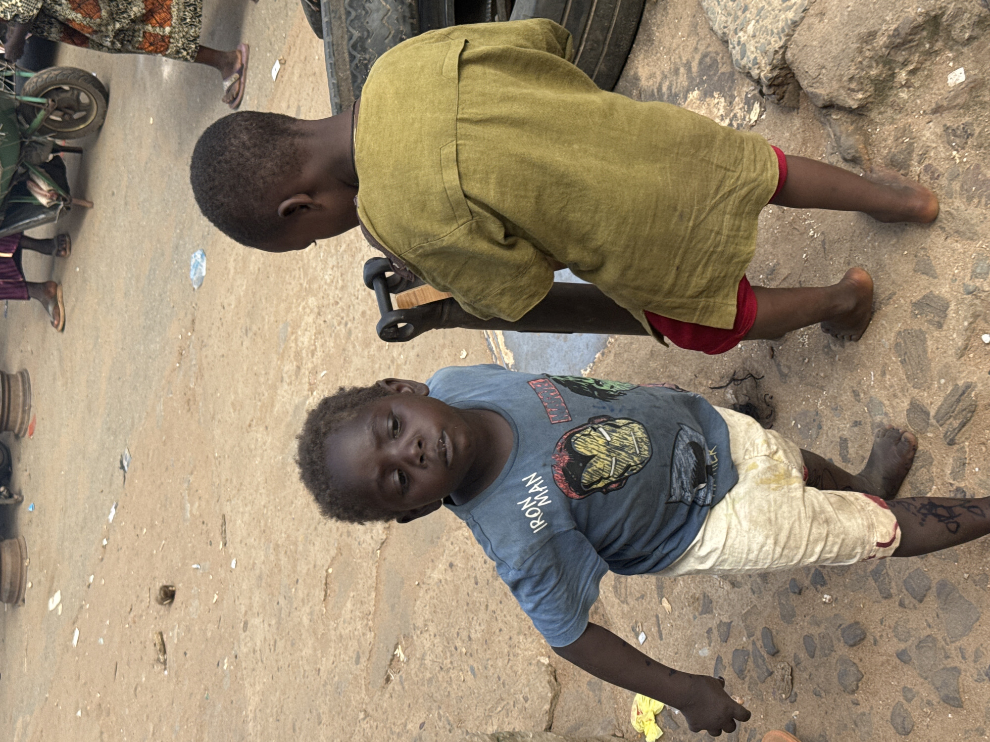 Two young Black children are standing on a dusty, uneven dirt and stone ground in an outdoor market area in Nkpor, Nigeria. The child on the left, facing forward, is wearing a blue t-shirt with "IRON MAN" printed on it and graphics of Iron Man's helmet. They are also wearing light-colored shorts. The child on the right, facing away from the camera, is wearing a green tunic and red shorts. They are interacting with a dark metal object with a wooden handle. In the background, there are discarded tires, some debris on the ground, and parts of what appears to be a wheelbarrow or cart. The lighting suggests it is daytime.