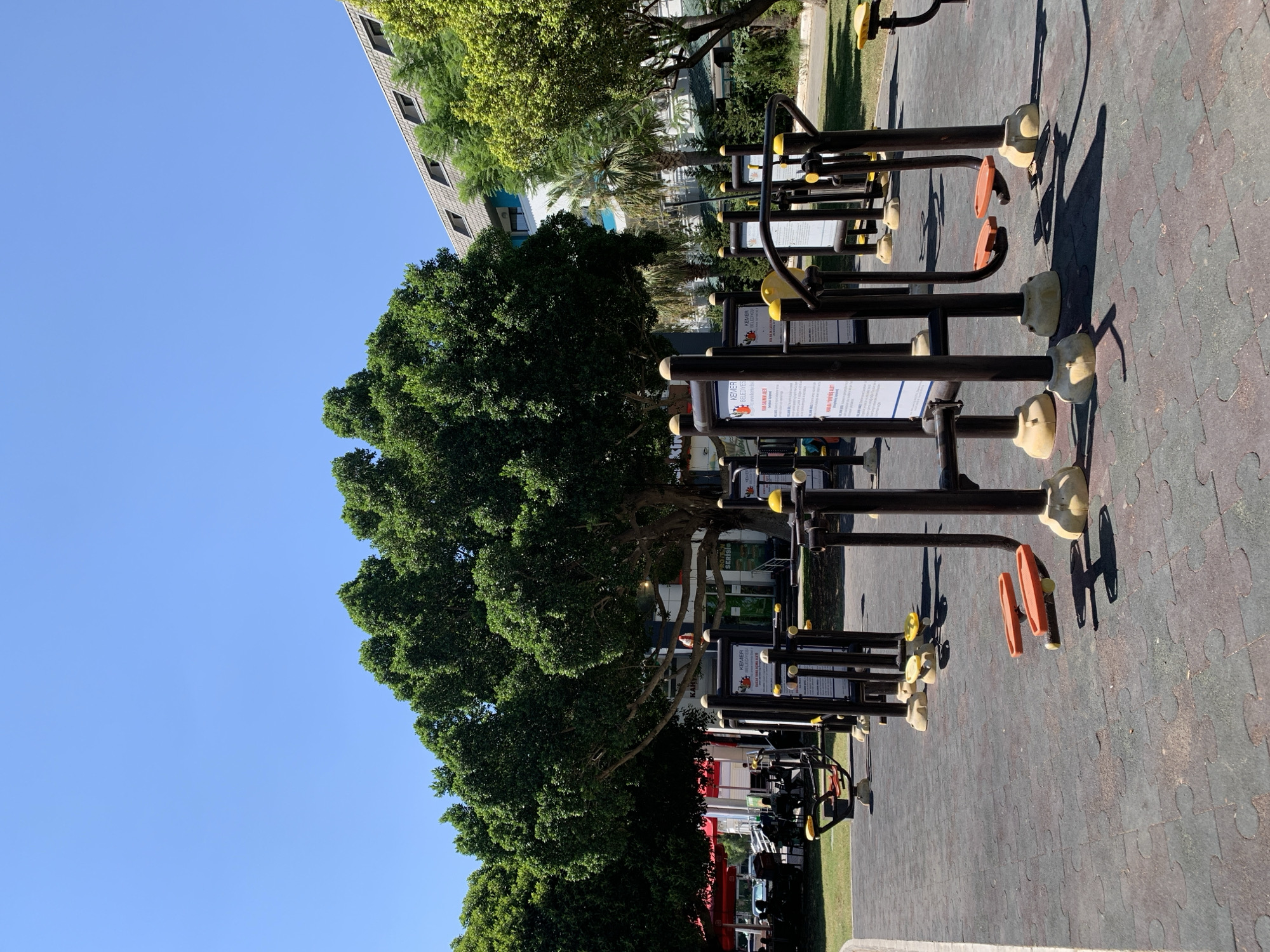 The image depicts an outdoor exercise area in Kemer, Turkey, bathed in bright sunlight under a clear blue sky. Several pieces of fitness equipment, primarily consisting of dark metal frames with orange and yellow accents, are arranged on a textured dark grey ground surface. In the background, a large, lush green tree with a thick trunk dominates the scene, with a building featuring a grey, sloped roof and several windows visible behind it, partially obscured by more greenery. To the left of the main tree, another large tree shades a seating area with red umbrellas and tables where several people are seated, suggesting a cafe or restaurant. The overall environment appears to be a public space, possibly a park or a recreational area within a resort.