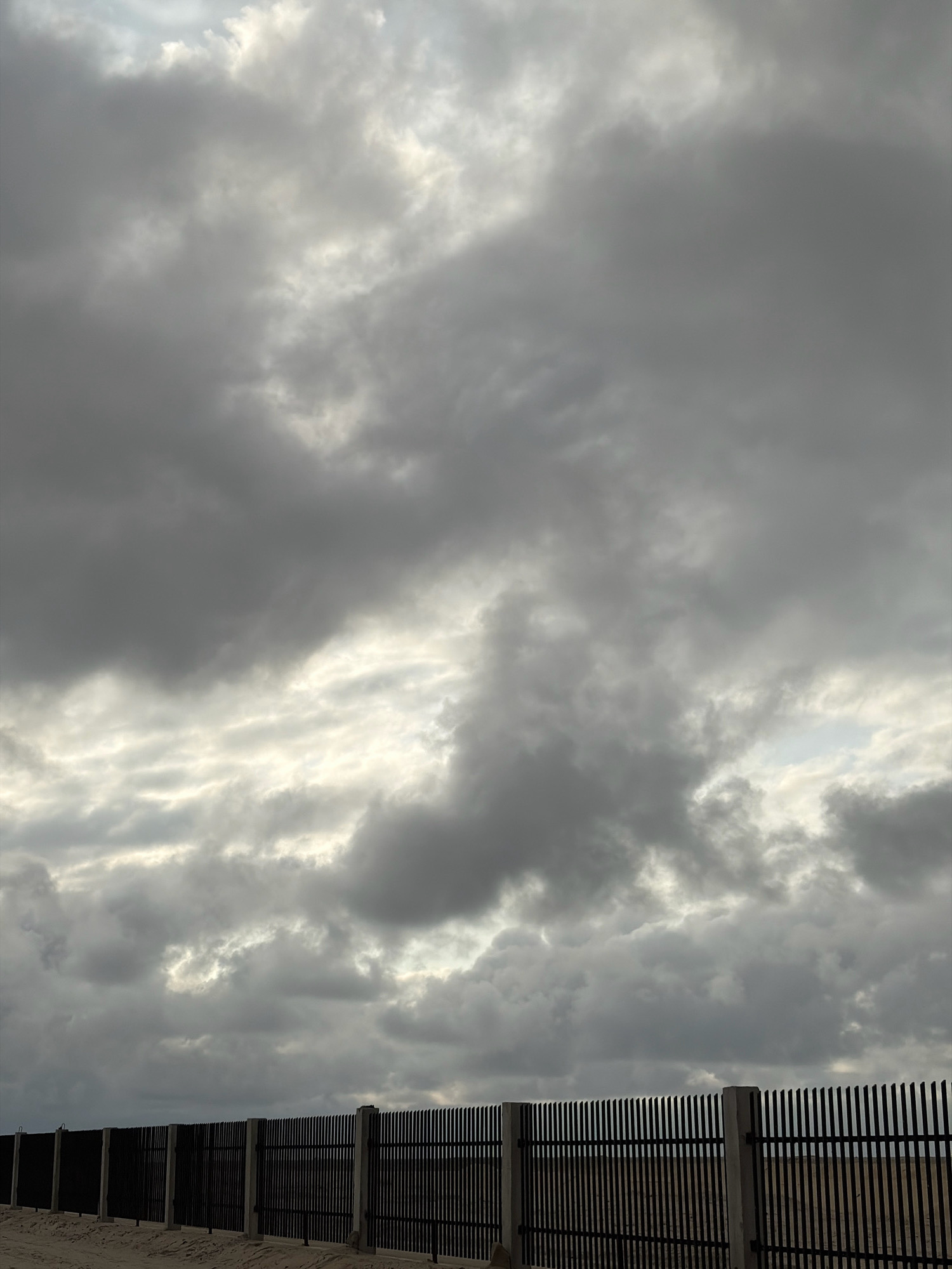 The image depicts a segment of a black metal fence with concrete posts, bordering a sandy area. The fence runs horizontally across the lower portion of the frame. Above the fence, a heavily overcast sky dominates the scene, filled with dark gray and lighter gray clouds. The lighting suggests a diffused, possibly pre-storm or post-storm, atmospheric condition. There are no people visible in the image. The location is identified as Alaguntan, Nigeria.