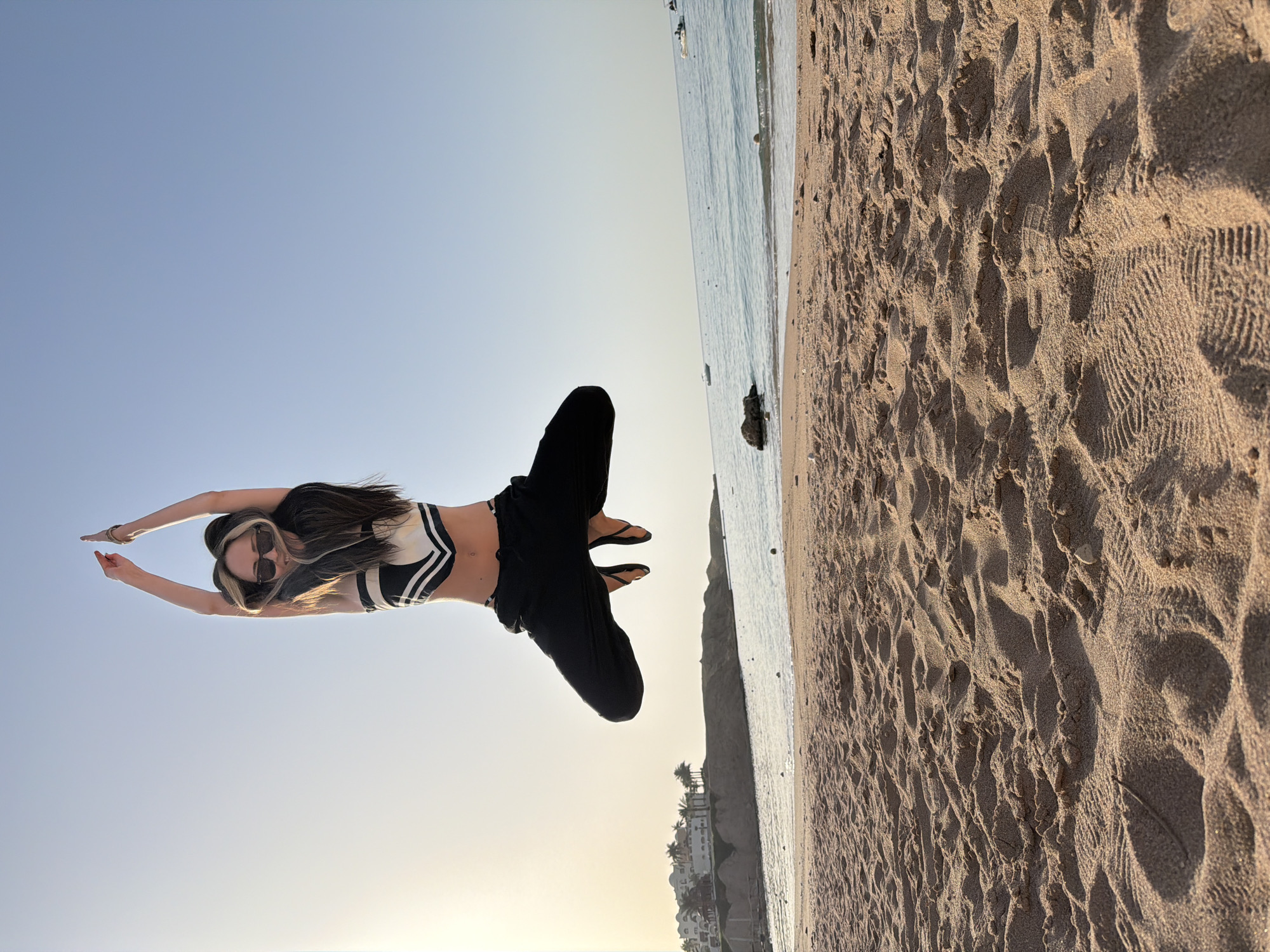A young woman is suspended in mid-air above a sandy beach. She is wearing a black and white striped top, black pants, and sunglasses. Her arms are extended outwards. In the background is a beach with visible footprints, the ocean, and some buildings. The location appears to be Sharm El Sheikh, Egypt.