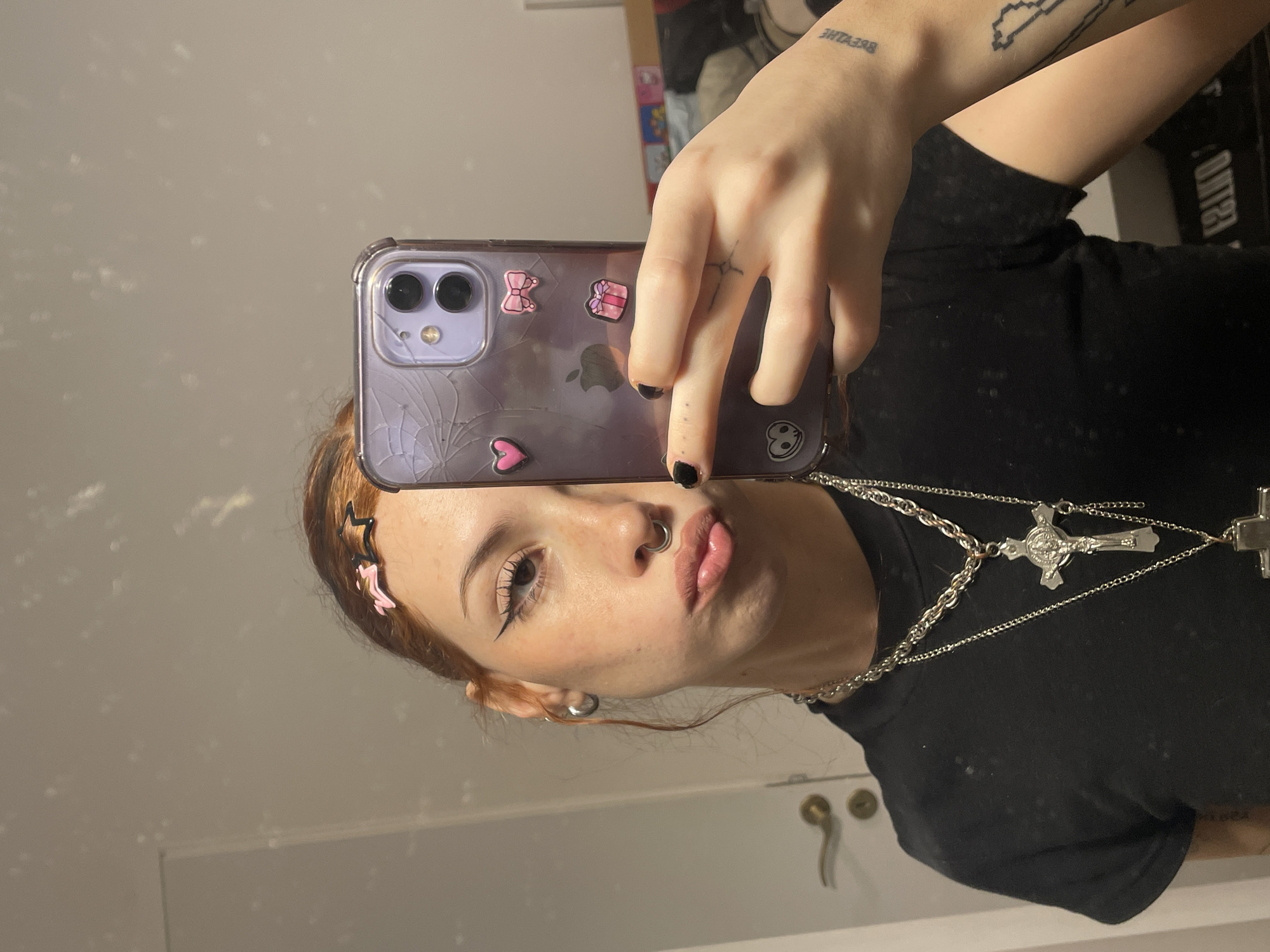 A young person is taking a selfie in a mirror. The person is wearing a black t-shirt, a silver necklace with cross pendants, and has a nose ring. They have light brown hair and winged eyeliner. Their phone has a cracked purple case with stickers. The background includes a white door with silver hardware and part of a closet with items inside. There is no visual information to confirm the location in Buenos Aires, Argentina.
