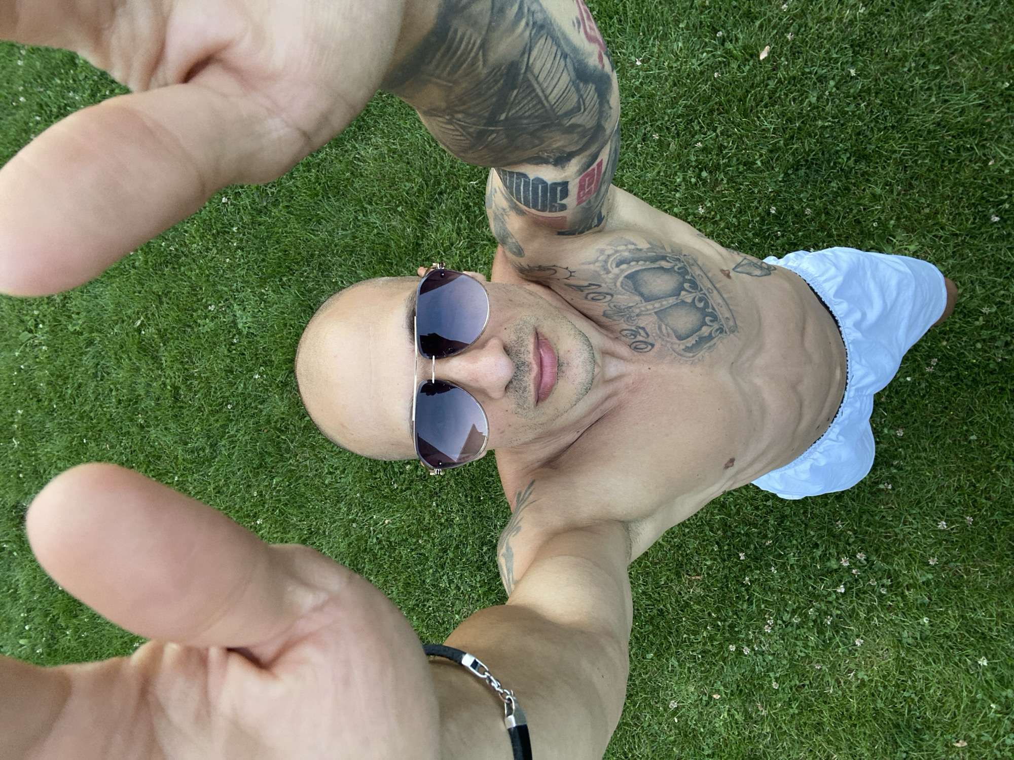 A bald, shirtless man with sunglasses and tattoos on his chest and arms is lying on a grassy lawn. His hands are raised towards the camera, obscuring his face. The man is wearing white shorts. The background is a green lawn with scattered white clover flowers. The image appears to be a selfie taken outdoors. The location is not specified.