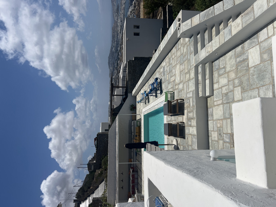 The image displays an outdoor, multi-level terraced property located in Tourlos, Greece. The scene is illuminated by direct sunlight under a blue sky with scattered white cumulus clouds.
In the midground, a rectangular swimming pool with clear turquoise water is central. Immediately adjacent to the pool, on a light-colored flagstone patio, are two dark-colored wooden sun loungers with striped cushions and a closed dark blue patio umbrella. Further along the pool's edge, four light-blue reclining sun chairs with white frames are arranged.
To the left of the pool, a covered outdoor lounge area features a white roof supported by beams, housing multiple red-topped tables and light-colored modular seating arrangements. The structures throughout the property are white-washed, indicative of Cycladic architecture, featuring thick walls and flat roofs. Flagstone paving covers the terraced surfaces, which are connected by sets of stone stairs.
The background presents an expansive view of the coastal town of Tourlos, characterized by densely packed white buildings cascading down hillsides towards a bay and the Aegean Sea. Utility poles and power lines are visible on the upper hillside. A dark-colored vehicle is discernible on a road high on a distant hill. No individuals, animals, or explicit actions are present within the frame.