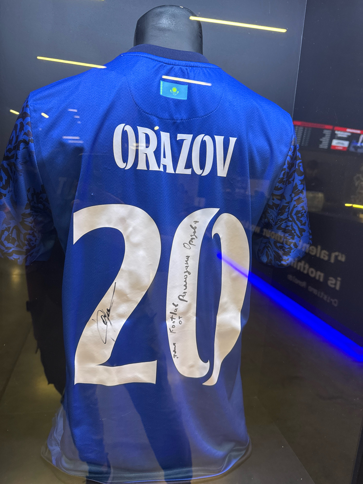 A blue football jersey with the name "ORAZOV" and the number "20" printed on the back is displayed on a mannequin. The jersey features a Kazakh flag emblem on the collar and a faded black pattern on the sleeves. A handwritten signature and inscription are present on the number "20". The jersey is displayed behind glass, reflecting neon blue lighting and a digital display in the background. This is likely at the Museum of Sport in Almaty, Kazakhstan, given the national team colors and prominent display of Kazakh sporting memorabilia.