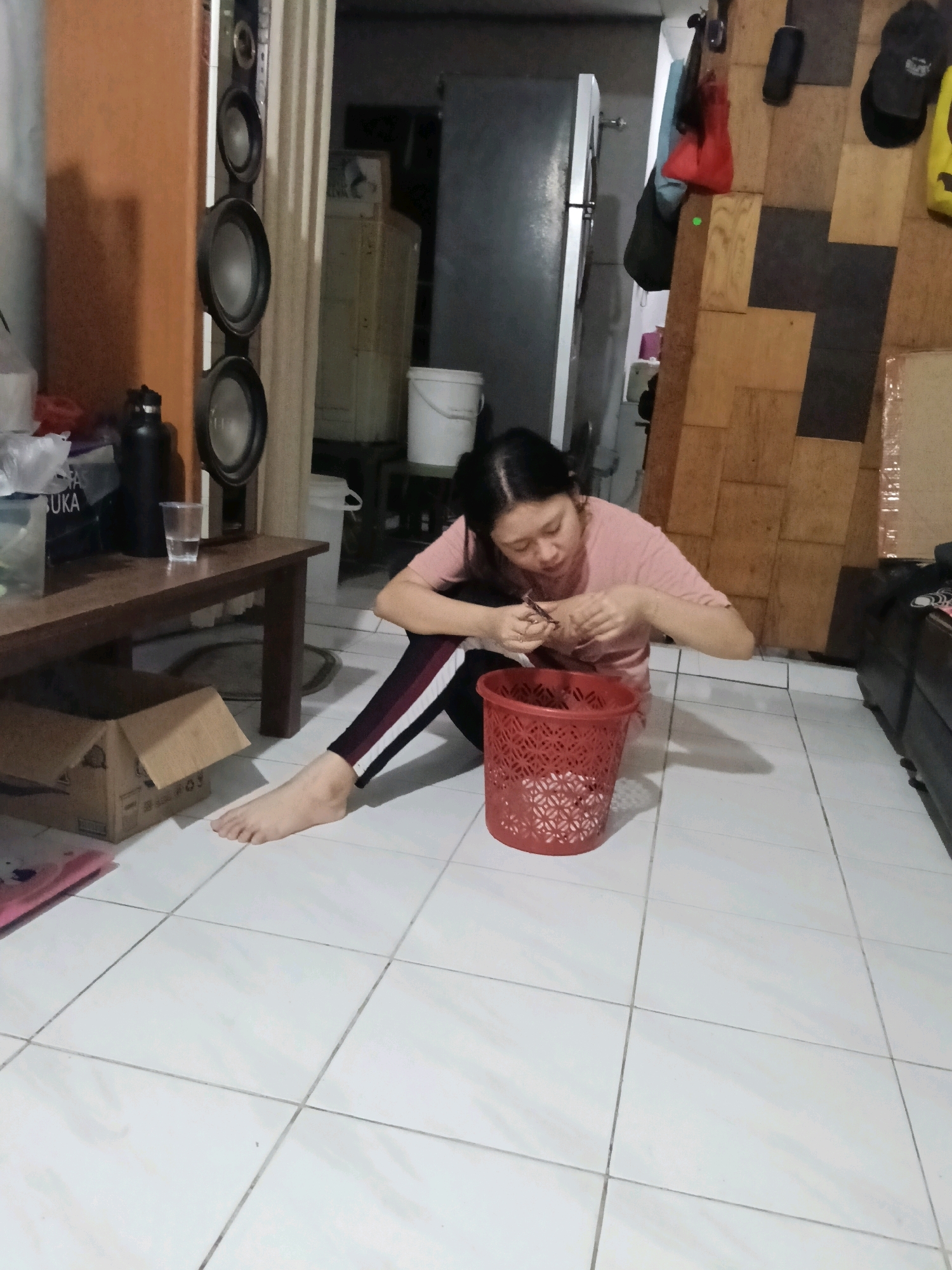 In what appears to be a residential dwelling in Jatimulya, Indonesia, a young woman is seated on the floor, engrossed in an activity. She is wearing a pink t-shirt and dark pants with white stripes. Her body is partially obscured by a red, perforated plastic basket placed in front of her, containing some white material, possibly waste. The woman is holding a pair of scissors in her right hand, using them to work on something held in her left hand. Her left leg is extended forward, while her right leg is bent. The floor is tiled with white square tiles, and a dark shadow stretches across it from the left. To the left of the woman, a wooden table holds various items, including a black water bottle, a glass of water, and what appears to be a cardboard box. Behind the table, a tall, dark speaker cabinet is visible, and beyond that, a doorway leads to another room. The background also features stacked wooden panels and a partially visible white bucket. The overall lighting is dim, suggesting an indoor evening setting.