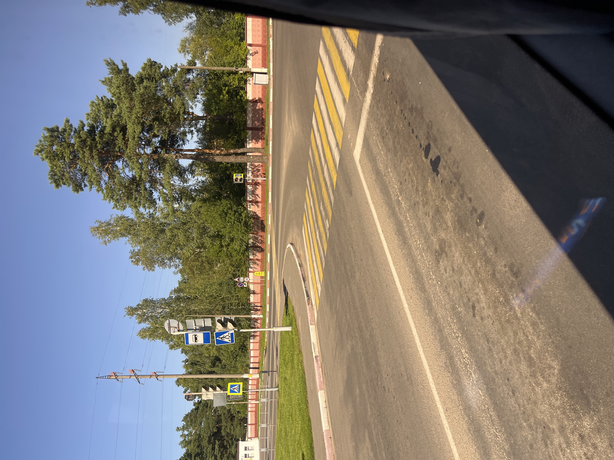 The image shows a street intersection in Pastavy, Belarus, under a clear blue sky. The foreground features a paved road with a prominent crosswalk marked by yellow and white stripes. A white line delineates a lane on the right side of the road, which is partially obscured by a dark shadow, possibly from a vehicle.
To the left of the crosswalk is a grassy area with a curb. Beyond the curb, a sidewalk is visible, bordered by a pink and white striped pattern. A traffic light pole with multiple signals stands near the intersection, displaying a red light. Adjacent to the traffic light is a blue sign with a white pedestrian symbol, indicating a crosswalk. Another blue sign above it depicts a bus, suggesting a bus stop. Further left, a yellow square sign with a white pedestrian symbol is also visible.
In the background, a long, red and white patterned wall runs along the edge of the road. Behind the wall, lush green trees form a dense backdrop. Tall pine trees and other deciduous trees are present. Electrical poles with overhead wires are also visible, contributing to the urban landscape. A small white building with windows is situated to the far left, near a grassy field. A speed limit sign displaying "50" is also visible on a pole near the wall. The overall scene depicts a typical daytime view of a quiet intersection in Pastavy.