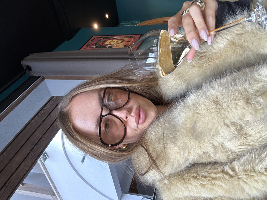 A female subject is depicted indoors in Courchevel, France, holding a stemmed glass containing a light-colored liquid, likely champagne. She has long, light brown hair and wears oversized, dark tortoiseshell aviator-style sunglasses. Her lips are slightly parted, and she wears a gold hoop earring on her visible left ear. She is dressed in a light beige or cream-colored faux fur coat. Her right hand, adorned with several gold rings, holds the glass, revealing light pink manicured nails. The background features an arched white wall and wooden ceiling elements to the left, suggesting a bright area or window. To the right, a dark gray curtain partially covers a teal-colored wall, on which a framed poster with reddish-yellow tones is mounted. A dark ceiling with a recessed light fixture is visible above.