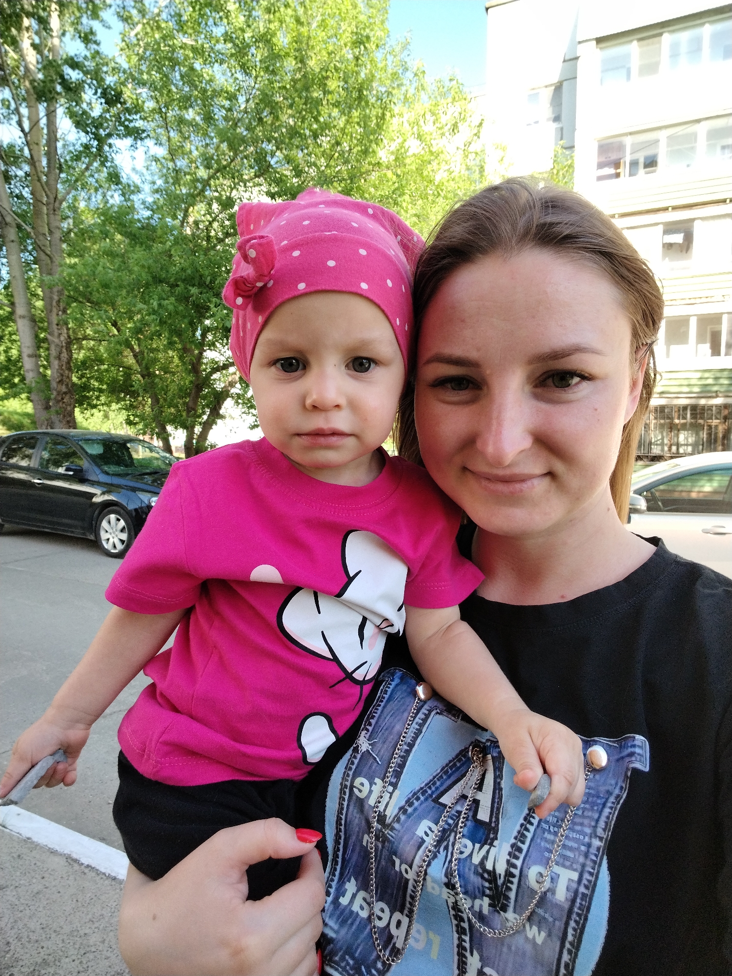 A woman holds a young child in her arms while standing outside a building in the city of Chaikovsky, Russia. The woman is wearing a black t-shirt. The child is wearing a pink, polka-dotted head covering, a pink shirt with a white cartoon design, and black bottoms. The child is holding a small grey object in one hand and a piece of blue denim fabric with a chain in the other. A black car is parked in the background. Trees and a building are visible in the background. The building has multiple windows and a light-colored facade.
