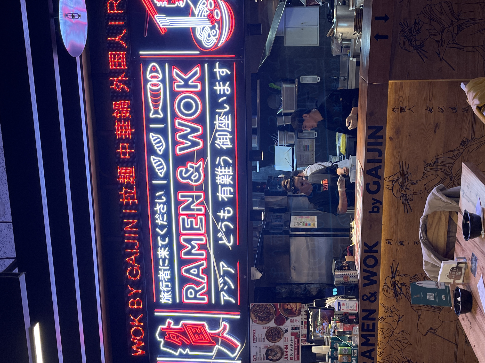 The image shows the exterior of a restaurant called "Ramen & Wok by Gaijin" located in Moscow, Russia. The restaurant name is prominently displayed in a neon sign with Japanese and English characters, with the name written in bright neon. A vertical electronic display shows "Wok by Gaijin" in English, followed by other Japanese characters. Inside the restaurant, two people are visible behind the counter, possibly chefs preparing food. Menus and product displays are visible below the counter. A wooden table is situated on the right side, with a QR code, napkins, and bowls. The walls behind the counter have wooden carvings of images and writing in Japanese. Wooden panels show "Ramen & Wok by Gaijin." Overall, the scene appears to be a bustling restaurant in Moscow with an Asian fusion concept.