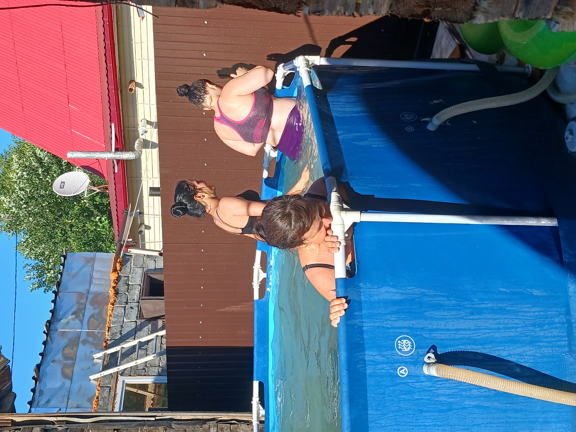 In an outdoor setting, two women are enjoying a swim in a blue, rectangular above-ground pool. The water is clear and inviting, reflecting the bright sunlight. One woman, in a purple bikini, is partially submerged, with her head tilted back and eyes closed. The other woman, wearing a black swimsuit, is leaning over the edge of the pool, appearing to reach for something in the water. Both women have their hair pulled back in buns.
The scene appears to be a backyard in Chik, Russia, with hints of residential structures in the background. A red-tiled roof and a brown wooden fence are visible, along with some greenery from trees. The time of day is clearly daytime, with strong sunlight casting shadows. The weather seems warm and pleasant, suitable for swimming.
Notable details include the inflatable nature of the pool, indicated by its white frame, and a flexible hose connected to a filter system at the bottom of the pool. There are also some circular symbols with wave-like patterns and a letter 'A' printed on the pool liner. The overall atmosphere suggests a relaxed and enjoyable summer activity.