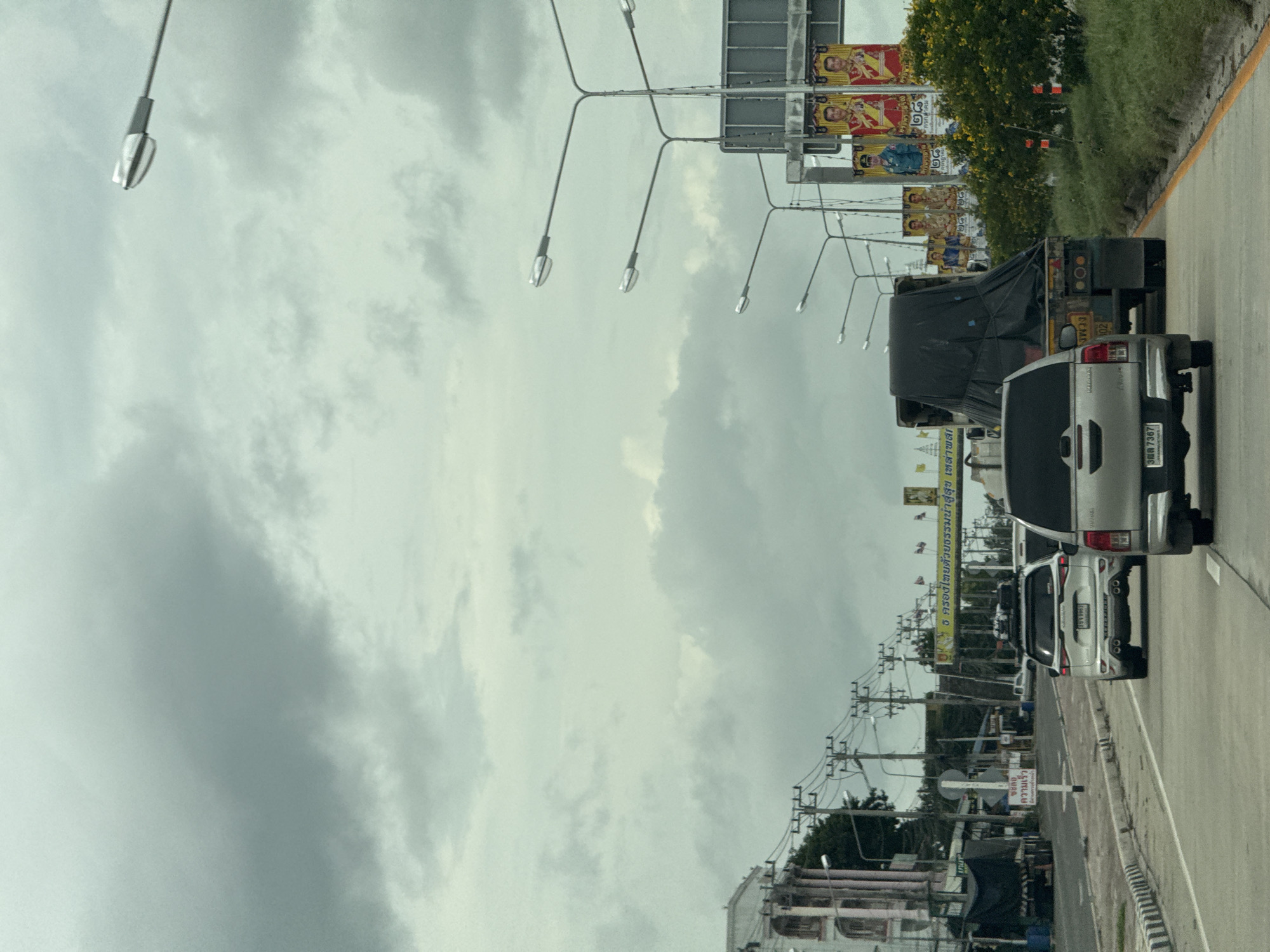 The image shows a highway in Ban Huai Yang, Thailand, on a cloudy day. Several vehicles, including a silver SUV and a truck with a black tarp, are traveling on the road. Buildings and utility poles are visible on the side of the road, along with several banners featuring portraits of royal figures. The sky is overcast with heavy clouds.