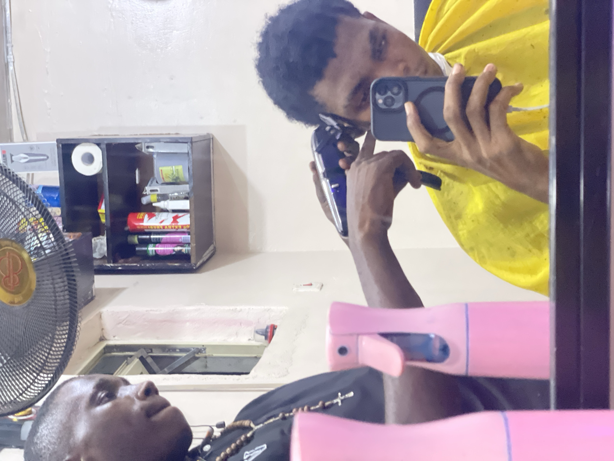 A selfie taken in a mirror shows two people, one lying down getting a haircut, with the barber visible in the reflection. The barber is holding a black phone and a hair clipper to trim the lying down man's hair. The barber is wearing a yellow cape. A black fan is visible to the left side of the image, and on the wall above the subject there is a black shelf with items stacked inside. A pink spray bottle sits in the foreground. The photo appears to be taken indoors and does not offer sufficient visual information to confirm whether this photo was taken in Ado, Nigeria.