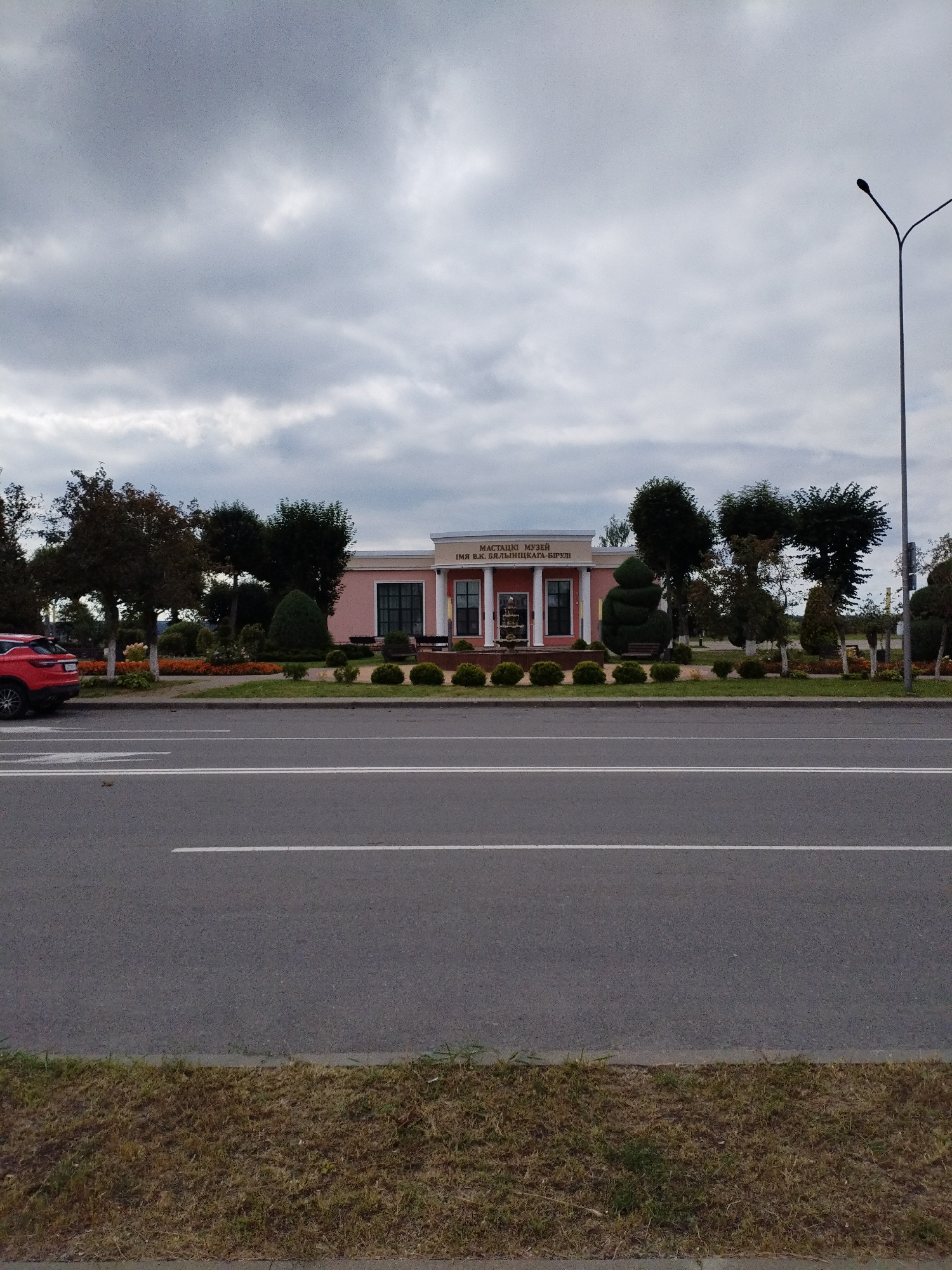 This is a full-frame, eye-level, outdoor, daytime shot of a building in Bialynichy, Belarus, under a cloudy sky. The building, identified by the sign "МАСТАЦКІ МУЗЕЙ ІМЯ В.К. БЯЛЫНІЦКАГА-БІРУЛІ," which translates to "Museum of Art named after V.K. Bialynicki-Birula," is a single-story structure with a light pink facade, white columns, and dark windows. It is situated behind a well-maintained lawn with sculpted bushes and flower beds. In the foreground, a multi-lane asphalt road with white lane markings runs horizontally across the frame. A red SUV is parked on the left side of the road, facing away from the camera. A streetlamp is visible on the right side of the image, rising into the overcast sky. The environment suggests a public space, likely an area with moderate traffic and pedestrian access to the museum.