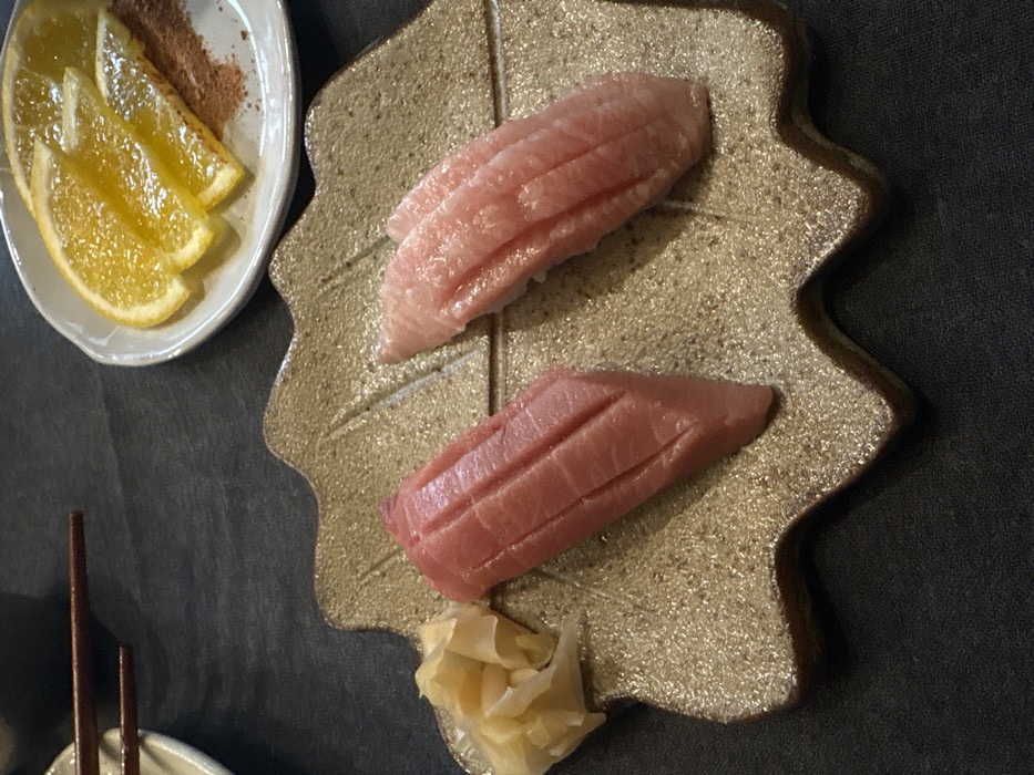 The image presents an overhead view of a Japanese meal artfully arranged on a dark tabletop. The central focus is a leaf-shaped ceramic plate holding two pieces of tuna sushi, likely nigiri. The top piece of sushi exhibits a pale pink hue with distinct marbling, characteristic of fatty tuna (otoro), while the bottom piece is a deeper red, suggesting medium-fatty tuna (chutoro). Both pieces of fish are scored with shallow cuts. Accompanying the sushi on the leaf plate is a small mound of pickled ginger (gari) at the bottom.
To the upper left, a small, light-colored, oval dish contains several bright orange slices, possibly for refreshment or as a palate cleanser. A small pile of reddish-brown powder, which could be chili powder, paprika, or cinnamon, rests beside the orange slices. A pair of dark wooden chopsticks is partially visible in the bottom left corner of the frame, resting on a small white holder.
The leaf-shaped serving plate has a textured, light brown or golden surface. The overall presentation indicates a thoughtful and refined dining experience, characteristic of a restaurant setting. The scene is devoid of people, visible text, or specific outdoor cues, but is situated within the context of Mexico City, Mexico, suggesting a vibrant culinary scene.