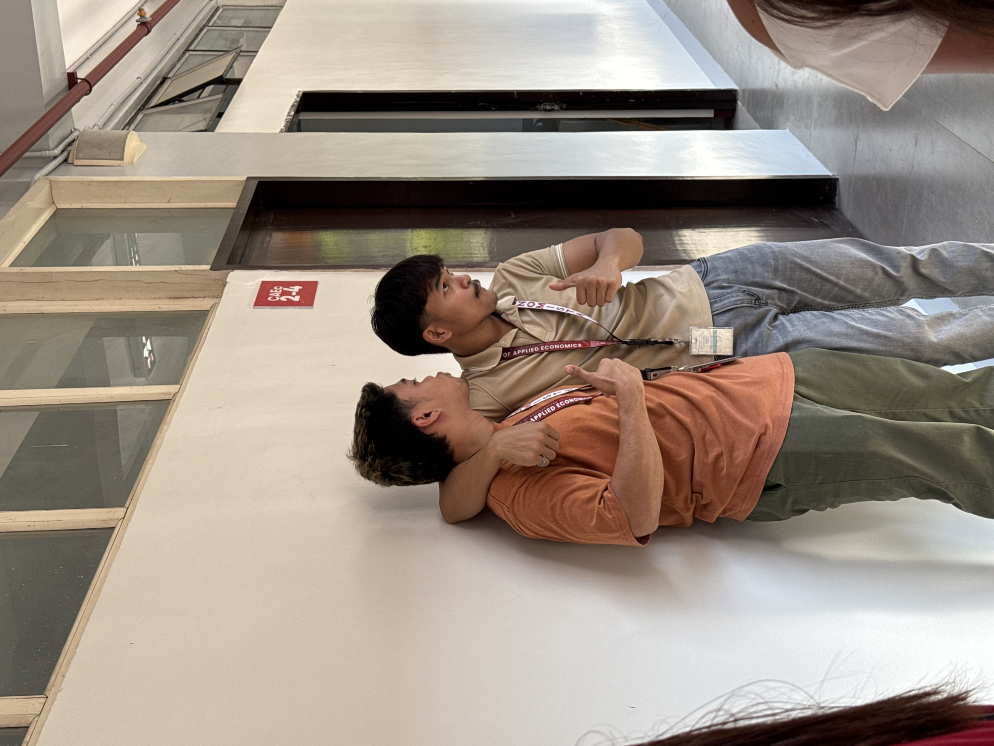 Two males are lying on a white floor indoors. The male on the bottom is wearing an orange t-shirt and green pants; the other is wearing a tan shirt and gray pants. Each is wearing a lanyard. The male on top points upward. A red sign with "CAEC 2-4" is on the white wall. Windows are visible in the background. The lanyards read "Department of Applied Economics." This suggests the scene is in an academic building in Davao City, Philippines, where the Department of Applied Economics is located.