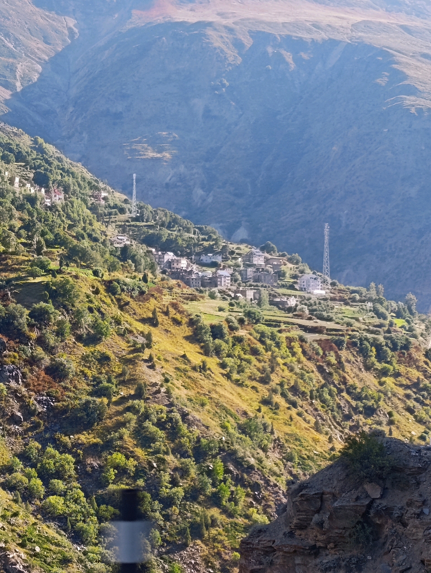 This is an aerial photograph of a steep mountainside with a village nestled into its slopes, located near Stingri, India. The village features a cluster of buildings with white roofs and grey walls, appearing to be constructed from stone. Terraced fields, covered in vegetation, are visible on the mountainside, indicating agricultural activity. Two tall, slender telecommunication towers are prominent features on the upper slopes, one to the left and another further to the right. The surrounding environment is rugged and mountainous, with vegetation ranging from sparse grasses and shrubs to more dense clusters of trees. The lighting suggests it is daytime, with sunlight casting shadows and highlighting the textures of the landscape. The overall impression is of a remote, high-altitude settlement.