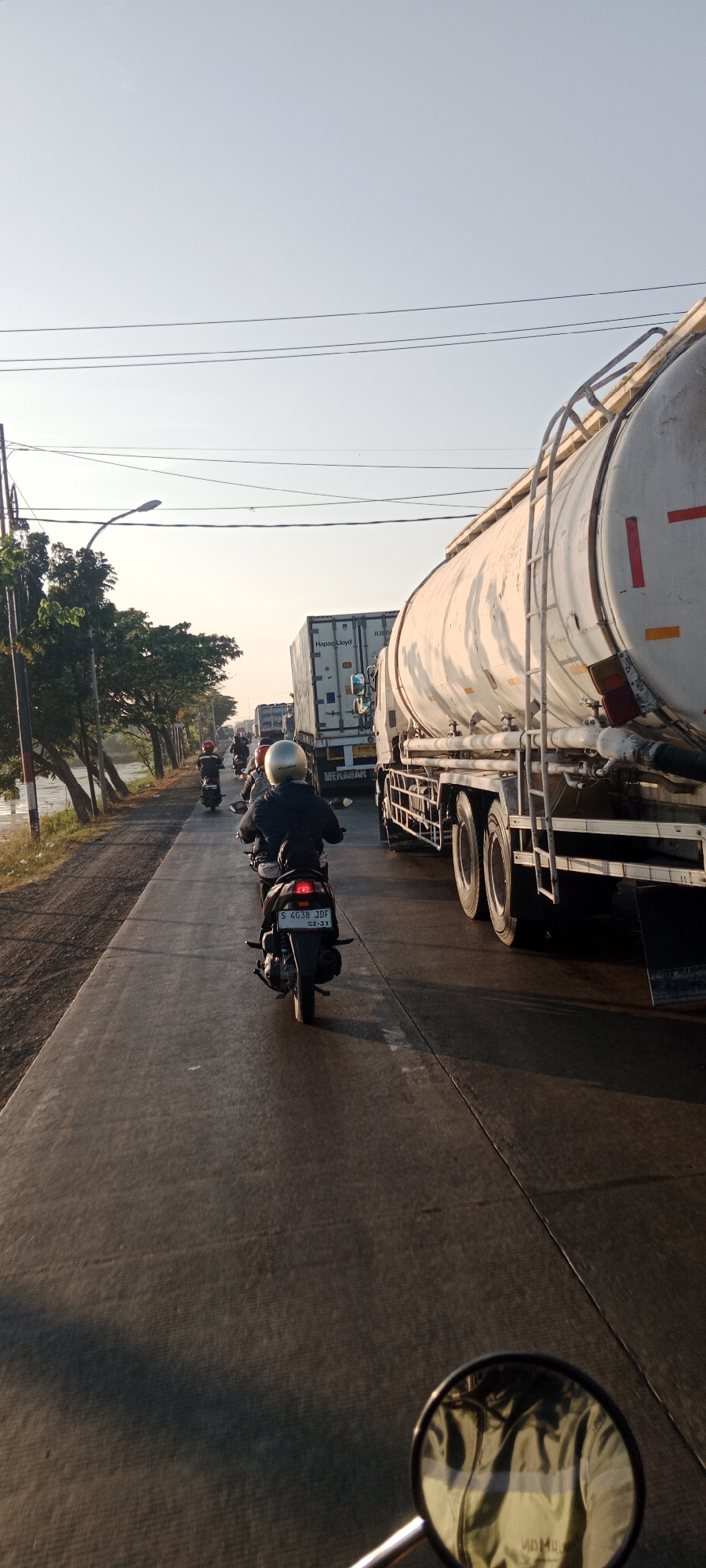 The image depicts a traffic jam on a road in Duduksampeyan, Indonesia, likely during the morning or late afternoon due to the low angle of the sun casting long shadows. In the foreground, a person is riding a motorcycle, with their reflection visible in the motorcycle's side mirror. The rider is wearing a helmet and a jacket. Ahead of this motorcycle, several other motorcycles are visible in the traffic jam. To the right of the road, large trucks are present, including a tanker truck and a container truck labeled "Hapag-Lloyd." Trees line the left side of the road, with a body of water beyond them. The sky is clear and light blue. The road surface is paved with asphalt and appears to have expansion joints. The license plate of the motorcycle in the foreground reads "S 4038 JDF."