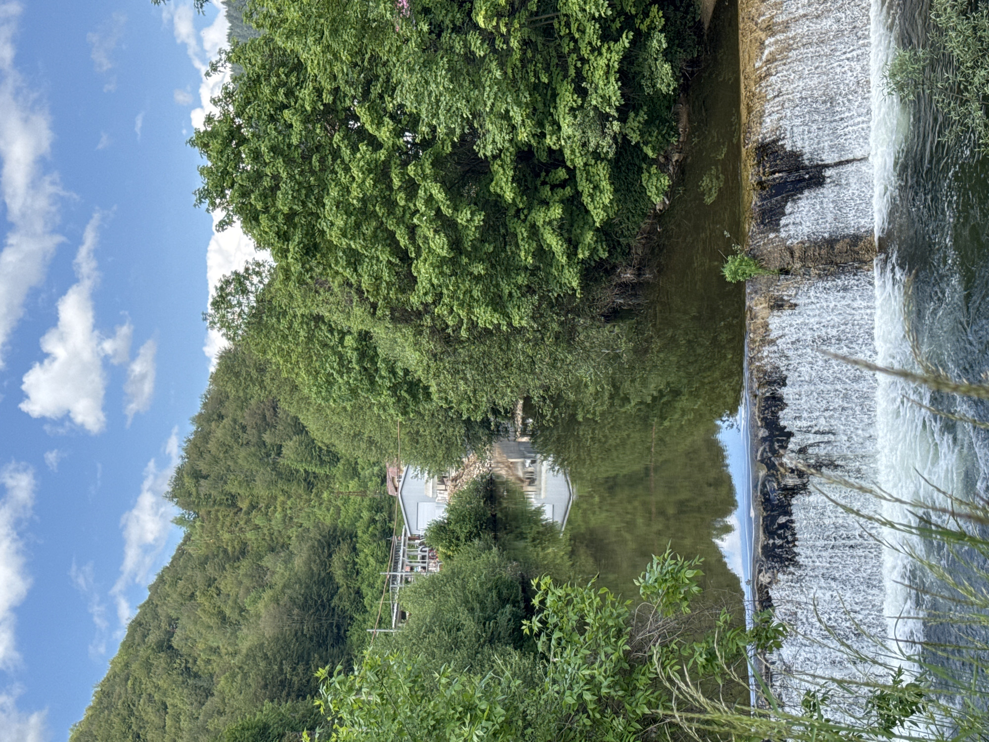 The image shows a waterfall in Rudozem, Bulgaria, with a lush green hillside and trees reflected in the calm water above. A white building is partially visible amidst the foliage, also reflected in the water. The sky is blue with scattered clouds. In the foreground, plants obscure some of the waterfall.