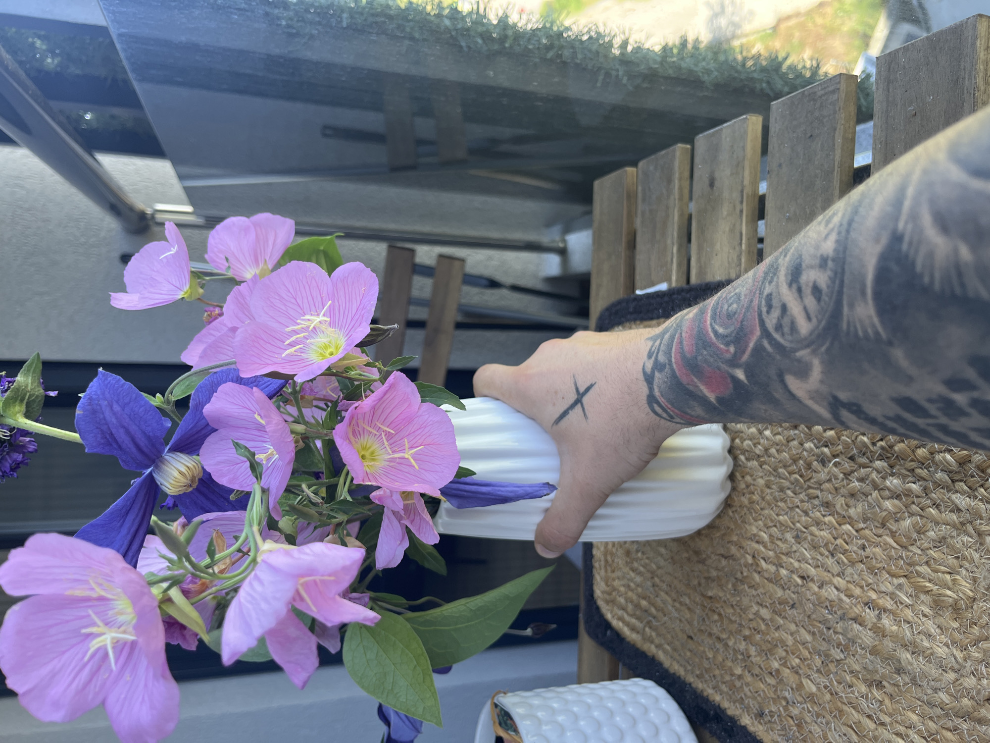 An adult male hand with a tattoo on the forearm is holding a white ribbed vase filled with pink and purple flowers. The vase is on a woven mat placed on a wooden table. The background shows a glass railing, wooden chairs, and greenery. The scene is outdoors, possibly on a balcony. The location is the city of Varna, Bulgaria.