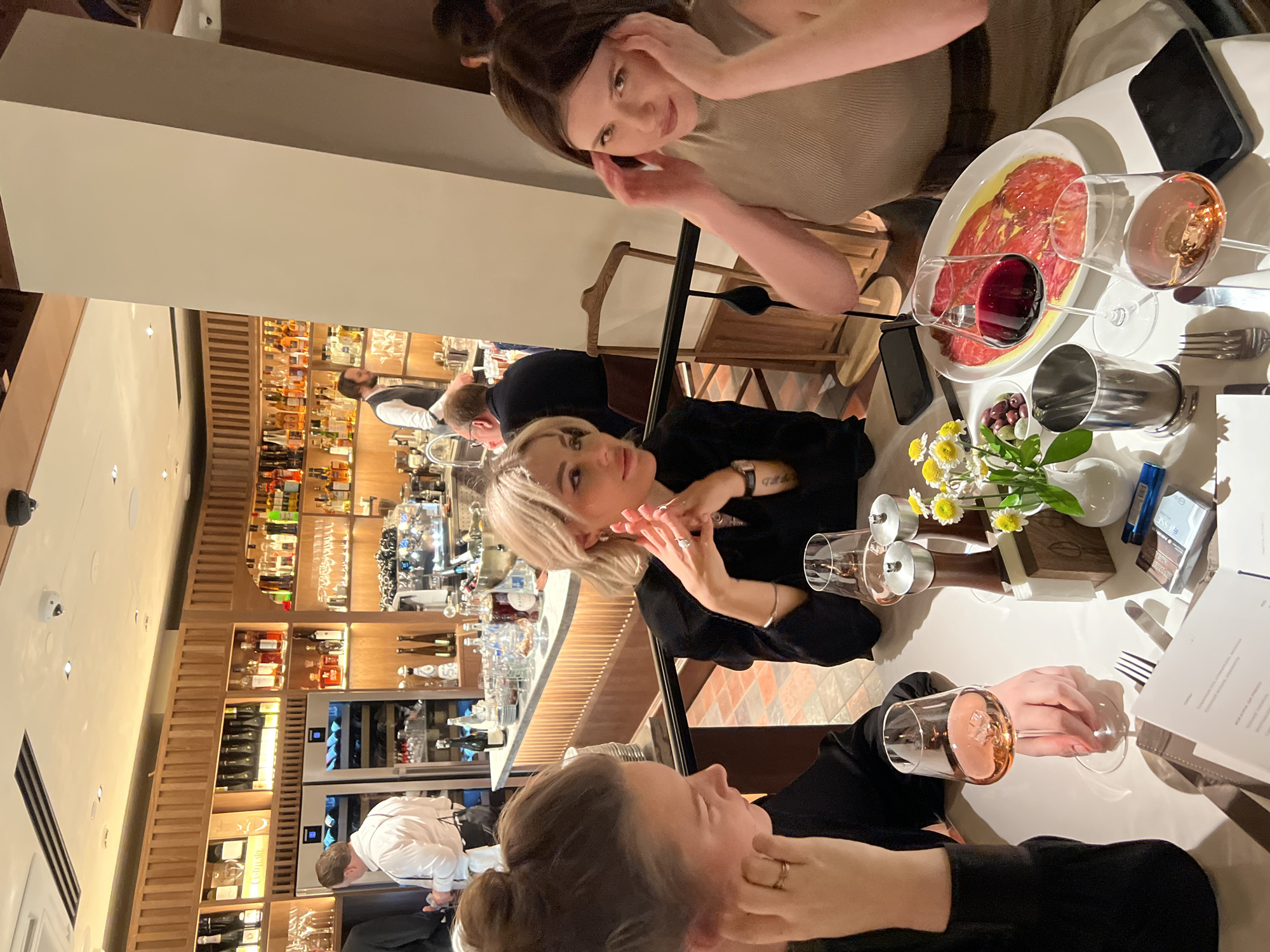 This image captures an indoor scene within an upscale restaurant or bar, illuminated by warm lighting, likely during the evening. Three women are seated around a white table in the foreground and midground, seemingly engaged in a social gathering.
In the upper part of the frame, a woman with dark brown hair looks directly towards the camera with a slight smile, her hands raised near her face. She is wearing a ribbed, light-colored top. Beside her on the table, a platter of thinly sliced red meat (possibly carpaccio) is presented, accompanied by a glass of red wine and an empty glass. A mobile phone is also placed near the edge of the table.
Towards the center, a woman with blonde, chin-length hair is visible, wearing a black long-sleeved top. She is looking to her left, with her hands clasped together, and a subtle tattoo is visible on her wrist. On the table in front of her are a small white vase containing white and yellow flowers, a dish of olives, and various other table settings including glasses, cutlery, and a white menu or pamphlet. A small blue box with the word "ESSENCE" printed on it is also present.
The lower portion of the image shows the third woman, viewed from the side, with her brown hair styled in a bun. She is also dressed in a black long-sleeved top, resting her hand near her chin, and holds a glass containing rosé wine.
The background showcases a meticulously organized bar area. Numerous bottles of various alcoholic beverages are neatly arranged on wooden shelves, complemented by built-in wine coolers. Two male bar staff members are visible behind the bar, actively working or conversing. A partially visible label on a wine bottle reads "Cantine". The overall atmosphere suggests a relaxed and sophisticated dining or drinking experience.