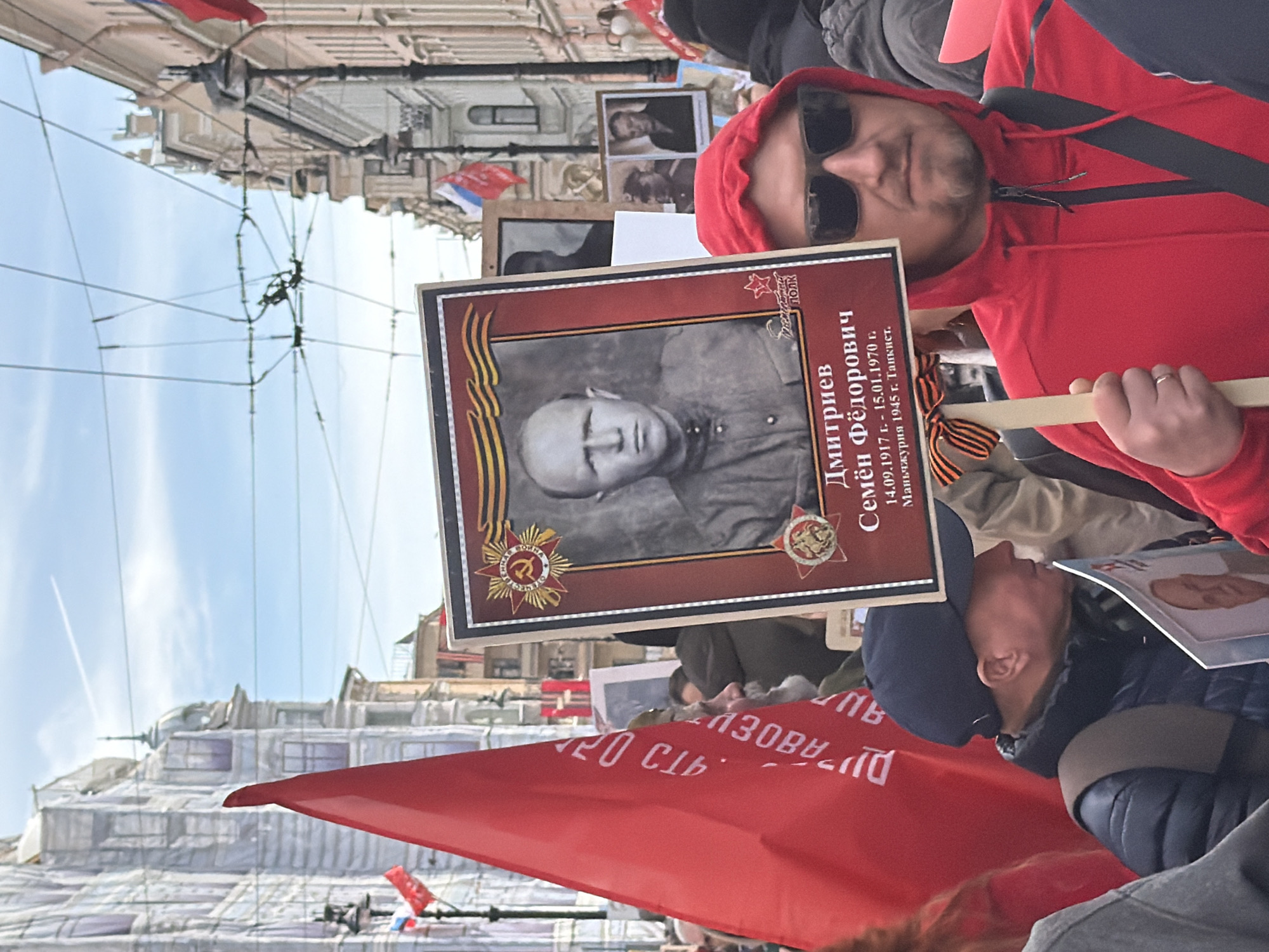 The image depicts a man in a red hoodie and sunglasses holding a framed portrait during a procession. The portrait is of a man in a military uniform, framed with a red border decorated with Soviet symbols, and bears the inscription "Дмитриев Семён Фёдорович 14.09.1917 г. - 15.01.1970 г. Маньчжурия 1945 г. Танкист." A Saint George ribbon is affixed to the portrait. Other portraits and a red flag with white Cyrillic text are visible in the background. The setting appears to be a street in Saint Petersburg, Russia, given the architectural style of the surrounding buildings and the presence of overhead tram lines. The sky is overcast. The context suggests participation in a Victory Day parade or similar commemorative event.