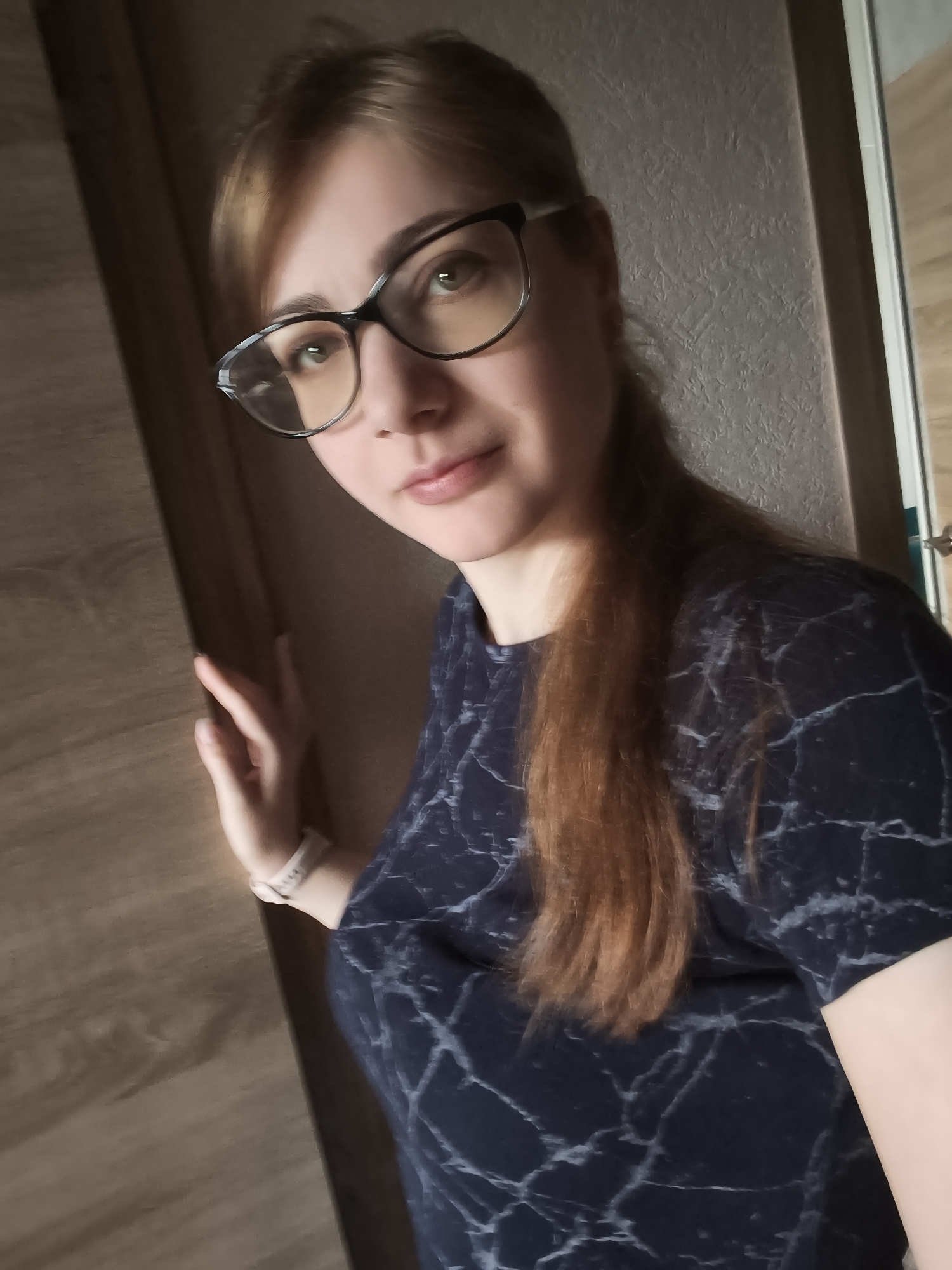 A medium shot shows a fair-skinned woman with auburn hair and glasses in a dark blue shirt with a white marble pattern. She is standing near a wooden door, her left hand resting on it. She is looking at the camera with a slight smile. The background consists of a textured wall and a doorway. The lighting suggests an indoor setting during the day. The image appears to be a selfie, as indicated by the angle and the woman's direct gaze at the camera. The scene is likely located in a residential environment, possibly a home in Podkletnoe, Russia, given the context.