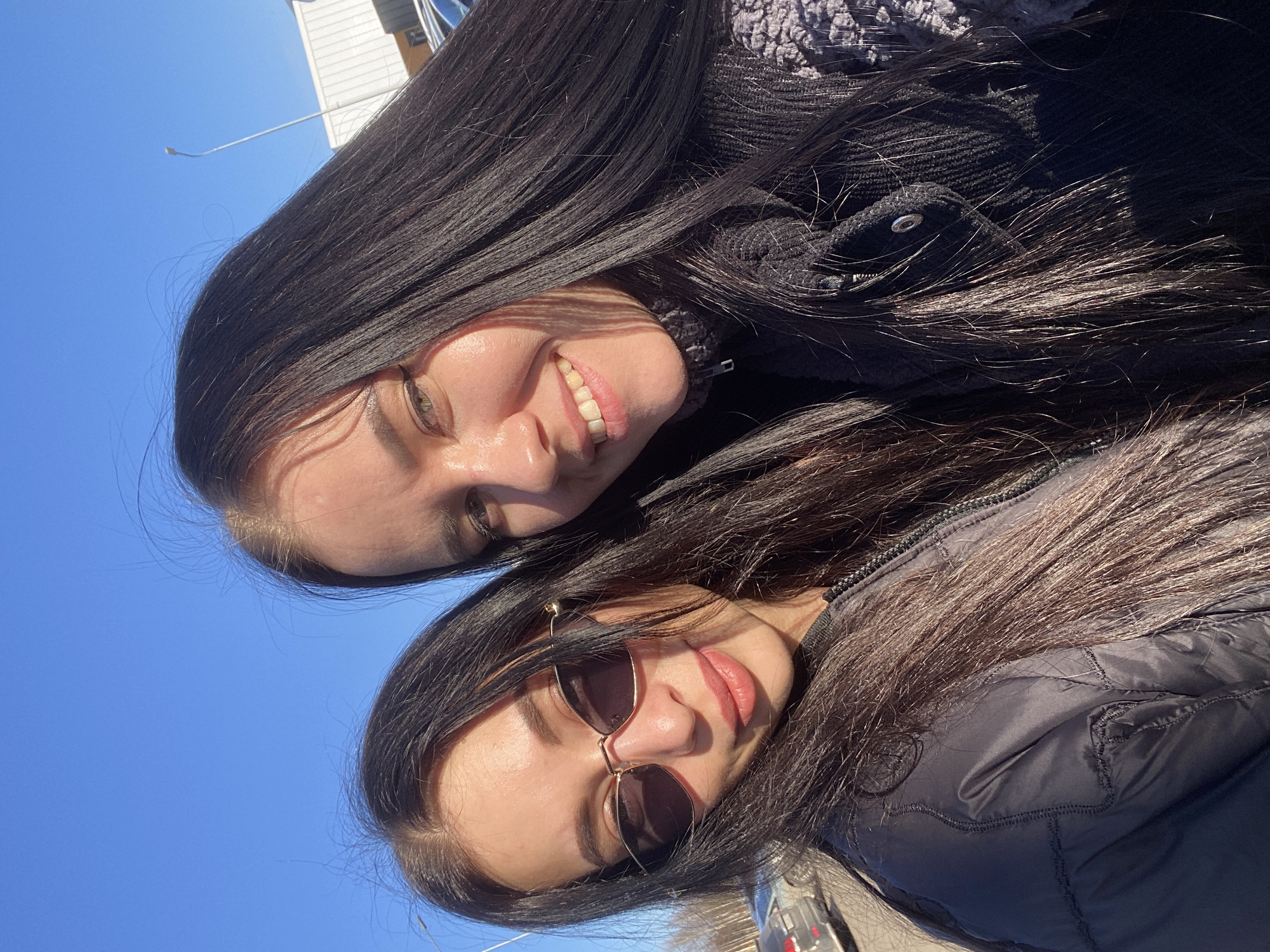 Two women with dark hair are posing for a selfie outdoors under a clear blue sky. The woman at the top is smiling, wearing a dark jacket over a textured sweater. The woman below is wearing sunglasses and a dark puffy jacket. Based on the sky and architectural elements visible in the top left, the location is likely an outdoor space. Because no specific landmarks or environmental details are visible, it is impossible to pinpoint the location more precisely than outdoors, or confirm the specific city of Votkinsk, Russia.