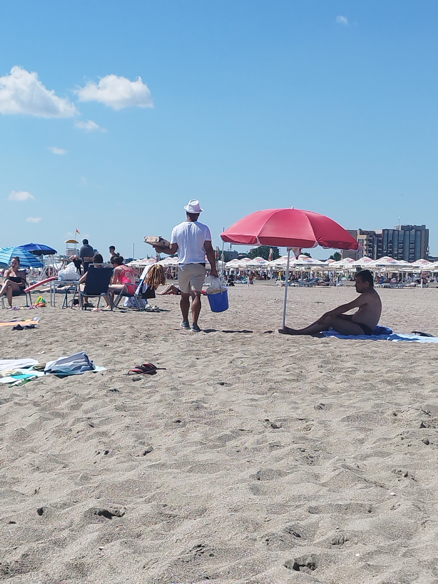 This is a medium shot of a beach on a sunny day, likely in Constanța, Romania, given the architecture visible in the background. In the foreground, the sandy beach is textured with ripples and footprints. A person is sitting under a red umbrella in the mid-ground, wearing black swim trunks and lying on a blue towel. To the left of the frame, a man wearing a white hat, white t-shirt, and beige shorts is walking away from the camera, carrying a flat object in his left hand and a blue bucket in his right. Further back, beachgoers are seated in chairs, and numerous beach umbrellas and lounge chairs are set up in rows. The sky is a clear blue with a few scattered white clouds. In the distance, buildings suggest an urban setting adjacent to the beach.
