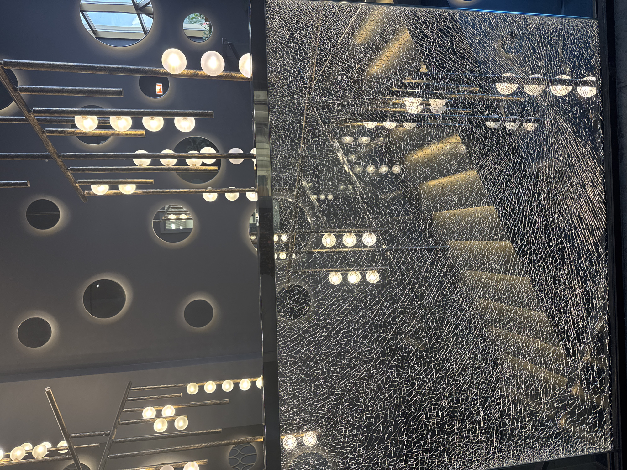 This is a photograph of a broken glass panel with a shattered pattern that reflects ambient lighting and circular mirrors embedded in a dark wall. The lighting fixtures are modern, with multiple suspended globes. The scene is interior, and the style suggests a commercial or hospitality space, possibly a bar or restaurant. There are no people visible in the image, and the focus is on the texture of the broken glass and the interplay of light and reflections. The specific location in Moscow, Russia, is not identifiable from the image itself.