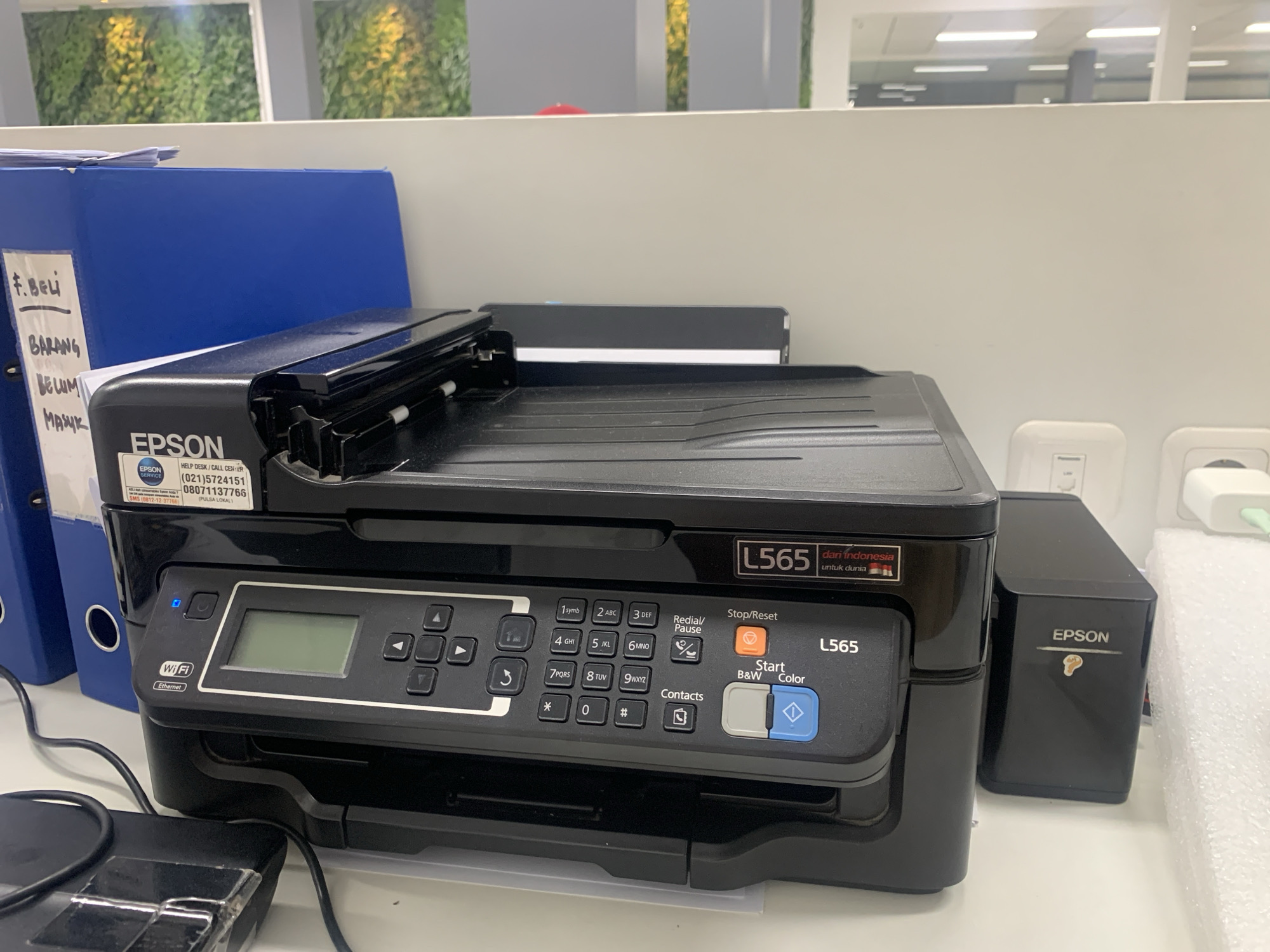 The image shows a black Epson L565 all-in-one printer situated on a white desk in an office environment. To the left of the printer is a blue binder with some handwritten text on a white sheet attached to it, and further left is another blue binder. To the right of the printer is a black Epson ink tank unit. On the wall behind the printer are some office lights and an abstract green wall covering. The scene is indoors, likely a typical office setting. The provided image does not contain any information that would allow for the determination of its specific location within Palembang, Indonesia.