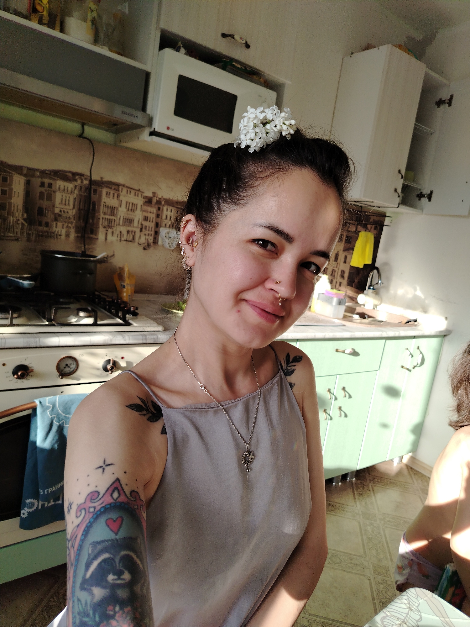A woman with dark hair pulled back, adorned with white flowers, smiles at the camera in a brightly lit kitchen. She has multiple piercings on her ears and nose. She wears a light gray tank top with a silver necklace. Tattoos are visible on her upper arm, including one featuring a raccoon inside a decorative frame with a red heart.
The kitchen has light green cabinets with a backsplash depicting a cityscape, possibly Venice. A gas stove with a black pot sits on the counter. A microwave is mounted above the stove. The flooring is tiled. Part of a child is visible in the lower right corner. Based on the location information and the general appearance, the scene is in an ordinary home in Noviy, Russia.