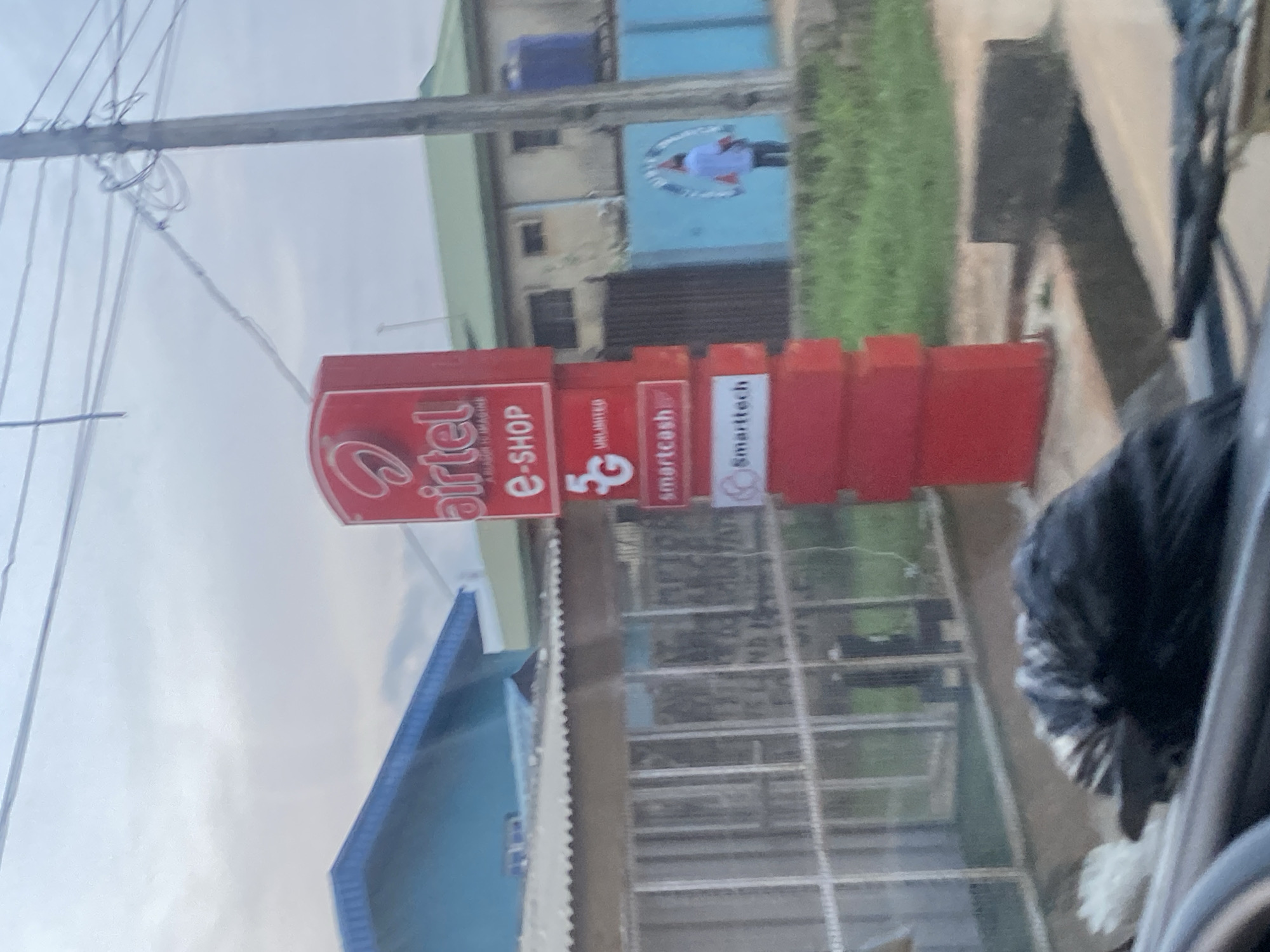 This media file captures a street scene in Ikorodu, Nigeria, likely during daylight hours given the bright sky. The primary subject is a red and white Airtel e-shop sign, prominently displayed on a vertical stand. The sign features the Airtel logo, "e-SHOP," and other branding elements including "UNLIMITED," "5G," "smartcash," and "Smarttech." Adjacent to the sign are stacked red rectangular boxes, possibly related to promotional materials or merchandise.
In the background, a building with a blue facade and windows is visible. Another building, partially obscured, has a light blue upper section and a darker lower section with windows. A person in a white shirt and dark pants can be seen in the distance, standing near a circular logo or graphic on the blue building.
The scene appears to be outdoors, with electrical wires strung overhead, suggesting an urban or semi-urban environment. The overall impression is of a commercial area, likely a street in a Nigerian town or city, with a focus on telecommunication services. The weather seems to be overcast or hazy, as indicated by the bright, diffused light and the appearance of the sky. A portion of a dark, possibly black, furry object is visible in the foreground on the right, which could be part of a vehicle or an animal.