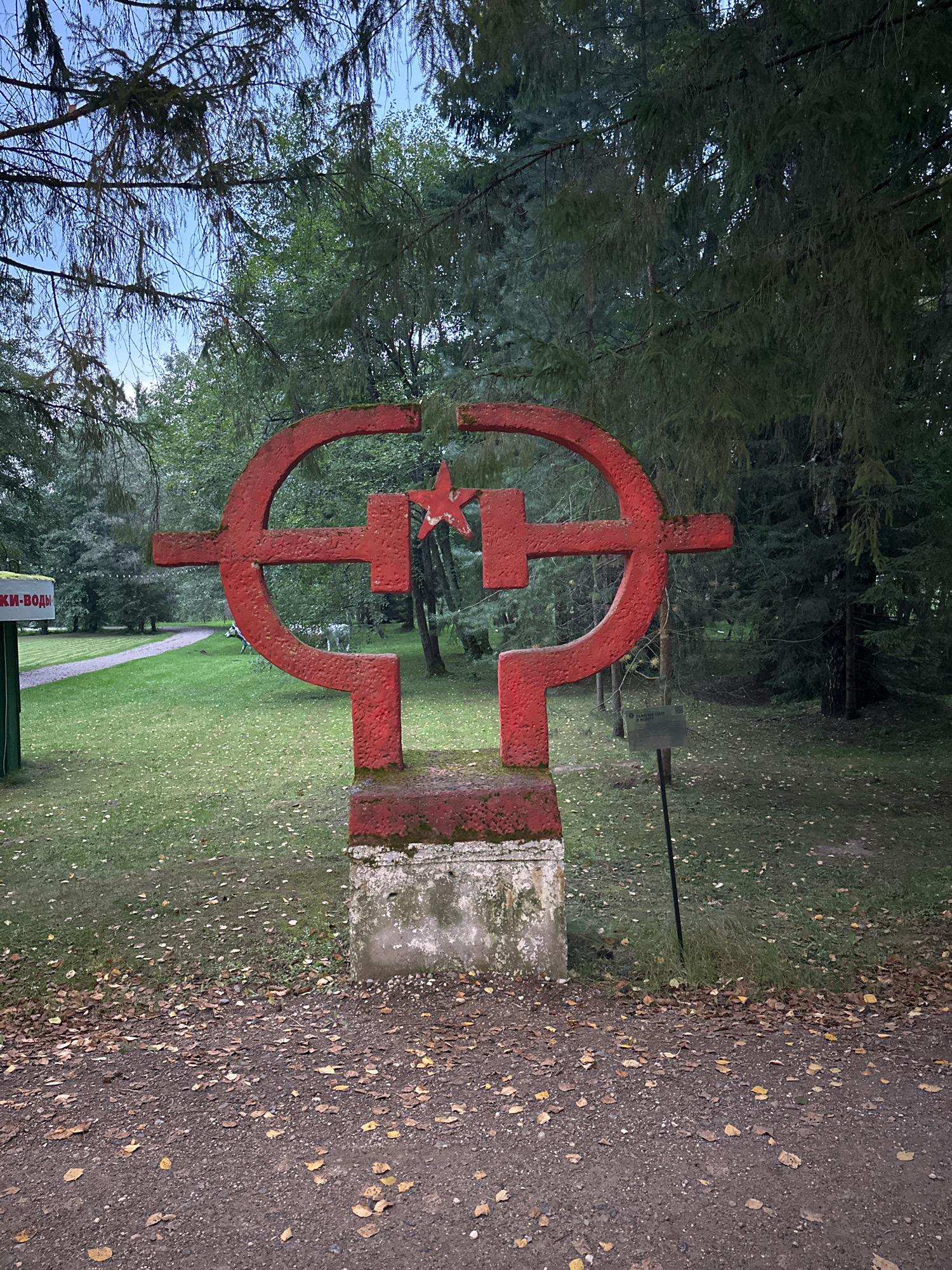 An outdoor, eye-level, medium shot captures a red hammer and sickle monument situated on a concrete base in Zyat'kovo, Russia. The monument, featuring a hammer crossed with a sickle and a red star at its center, is weathered with moss visible on its surface. It stands on a concrete pedestal that shows signs of age and damage. Surrounding the monument is a gravel path, strewn with fallen yellow and brown leaves, indicative of autumn. To the left, a green sign with Cyrillic text is partially visible, and further back, a paved path winds through a lush green lawn bordered by trees. The background is dominated by a dense forest of evergreen and deciduous trees, with a clear blue sky visible through the branches. A dark metal signpost stands to the right of the monument. The scene is set in daylight, though the lighting suggests it may be early morning or late afternoon. No people are visible in the frame.