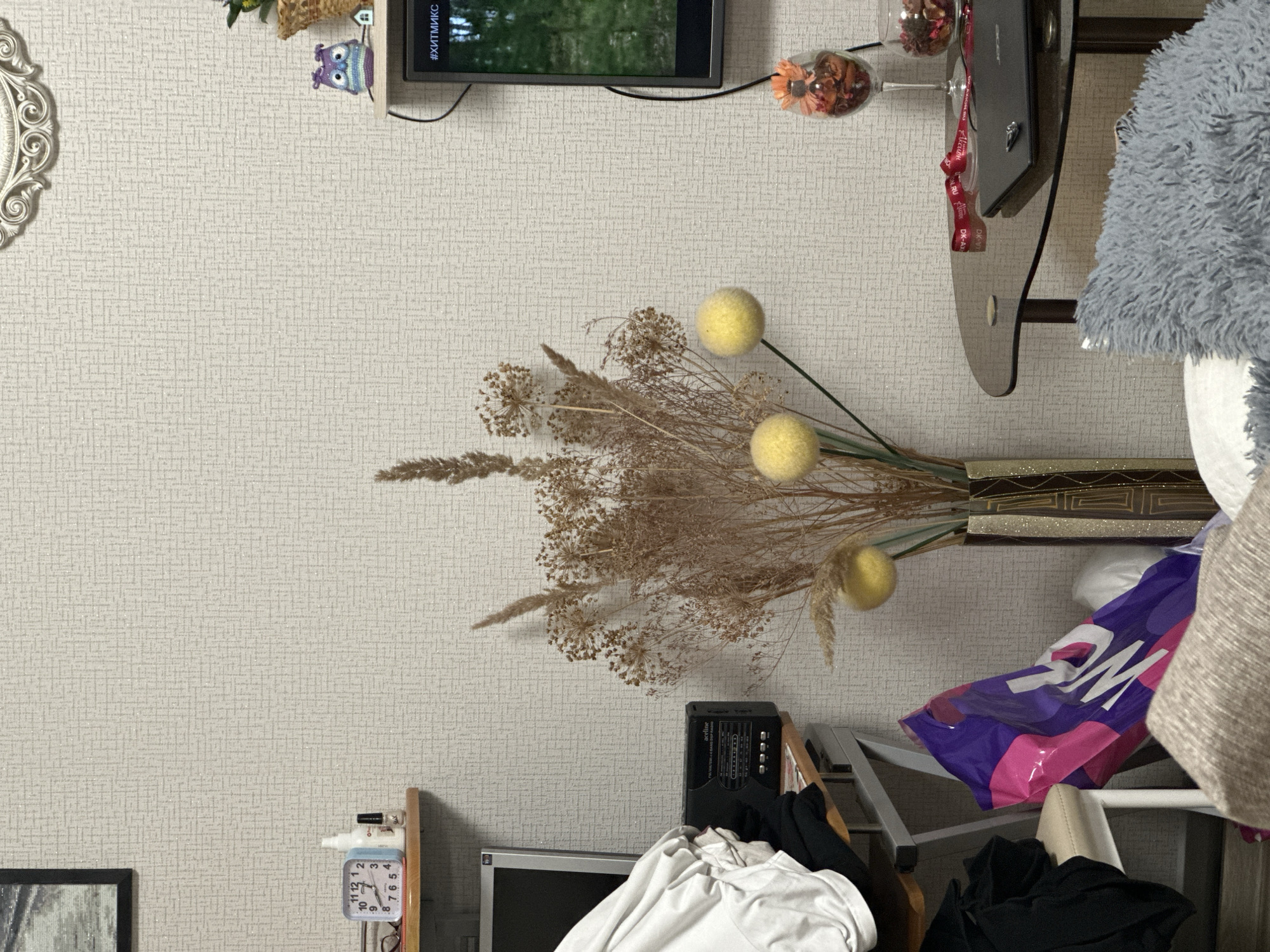 The scene presents an indoor setting, possibly a room within a residence, located in the city of Советско-Никольское, Russia. The decor is a mix of functional and decorative elements against a wall with a light-colored, textured wallpaper.
A television is mounted on the wall, and below it, a collection of decorative items including vases with dried floral arrangements and stemmed glassware. The flowers appear to be mostly dried grasses and pom-pom-like flowers in shades of brown and yellow.
A shelf or surface below supports miscellaneous items, including what appears to be a laptop or electronic device, and various textiles.
In the lower part of the frame, a smaller, older monitor and a small clock are visible amongst a pile of folded or crumpled clothing items. A radio is nearby. The assortment of clothing and other objects suggests a storage area or a space where items are casually placed. A shopping bag from Whataburger is present.