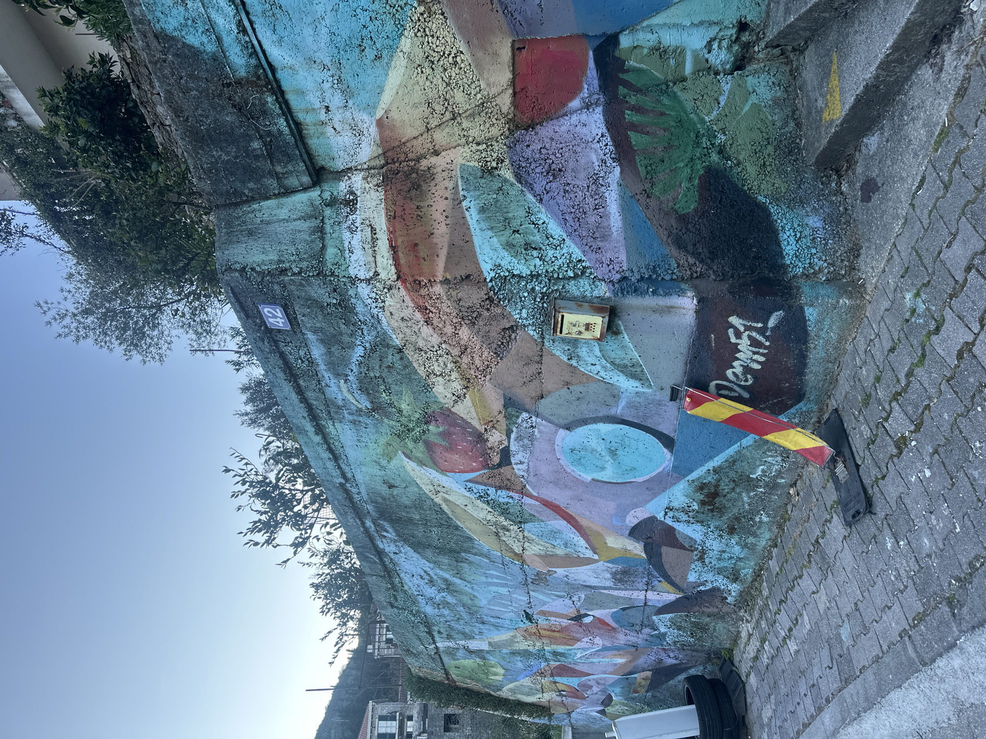 The image captures a section of a colorful, graffiti-covered retaining wall, likely in an urban environment. The wall appears to be made of concrete and is adorned with a vibrant mural composed of geometric shapes and organic forms in various colors. The mural exhibits signs of age and weathering. A small mailbox is affixed to the wall amidst the graffiti. A red and yellow striped barrier is positioned near the base of the wall, along the adjacent paved area. The paved area is constructed of interlocking bricks. Partial view of a vehicle with tires. Lush foliage and trees grow along the top edge of the wall. In the distance, glimpses of buildings and a hillside are visible. A blue sign displaying the number '42' is present. The clear sky indicates daylight. The location appears to be a public space in Budva, Montenegro, based on architectural elements and graffiti style common in European urban areas. The scene is static, with no visible people or dynamic actions.