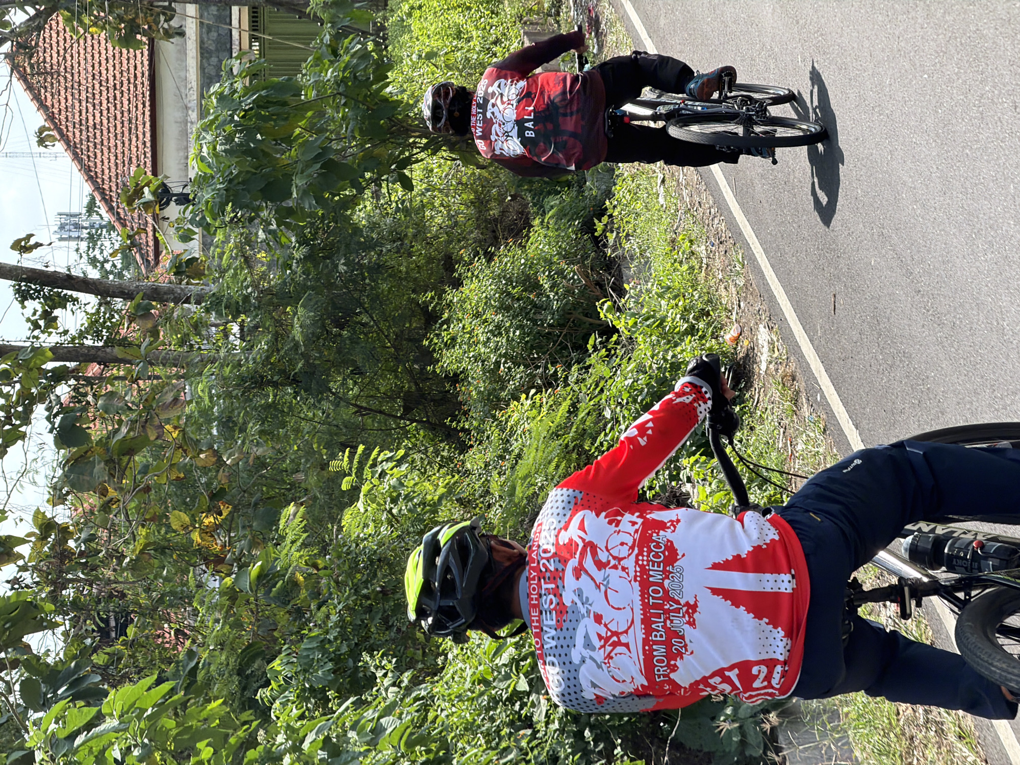 Two cyclists are riding on a paved road lined with lush green vegetation and trees. The cyclist in the foreground is wearing a lime green helmet and a red and white long-sleeved cycling jersey with graphics and text that reads "GO WEST 2025" and "FROM BALI TO MECCA 20 JULY 2025". The cyclist in the background is wearing a dark helmet and a red cycling jersey with "BALI" printed on the back. Behind the vegetation, there are glimpses of tiled roofs of buildings and communication towers. The sky is partly cloudy. The scene appears to be in a rural or semi-rural area of Kuta Selatan, Indonesia, suggested by the typical architecture and dense greenery.