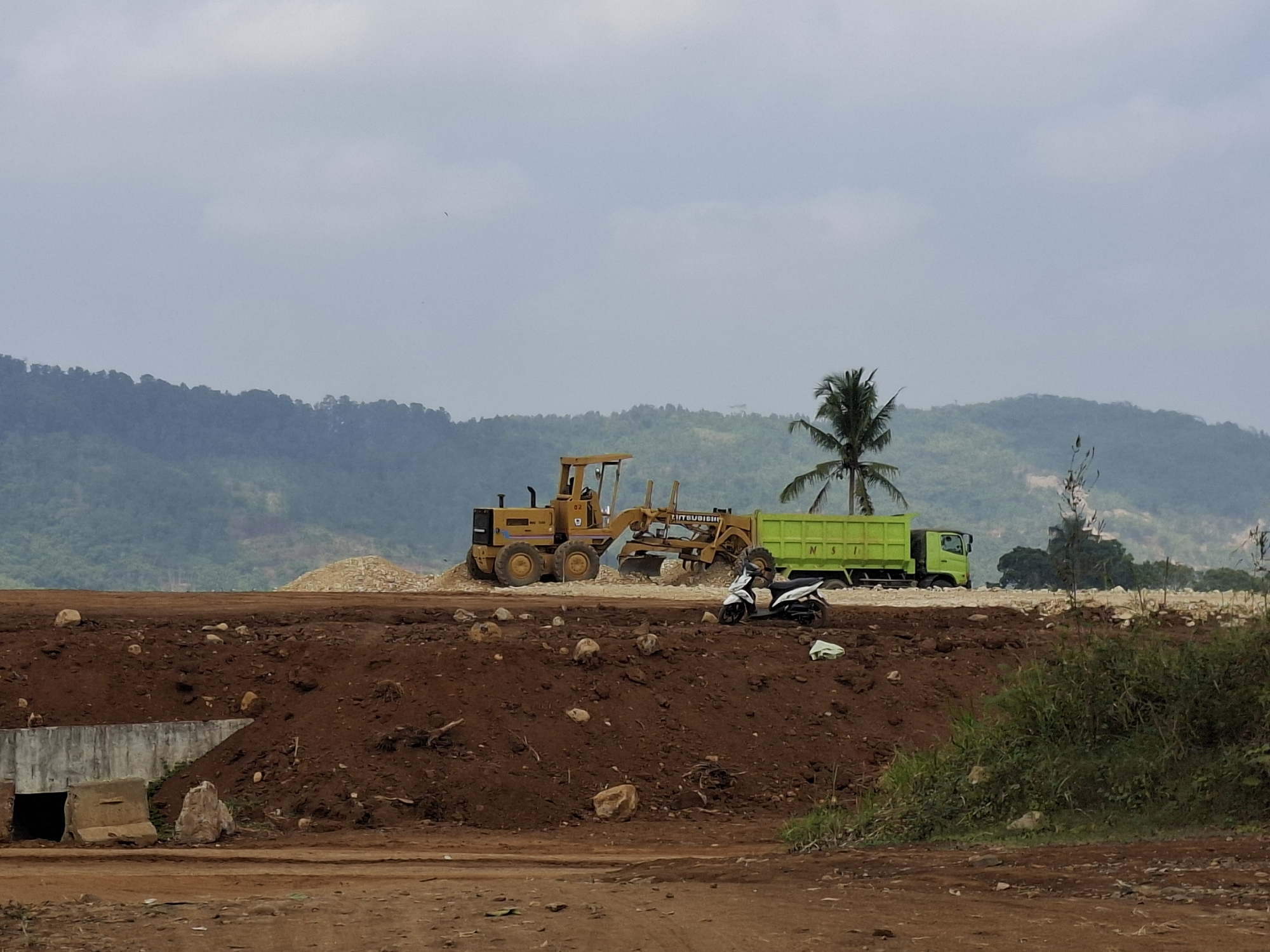 An outdoor, daytime scene depicts a construction site in Ciheulang Tonggoh, Indonesia. In the foreground, reddish-brown dirt forms a mound with scattered rocks. To the left, a concrete structure with a dark opening is partially visible. Further back, heavy machinery operates on a flat expanse of dirt. A yellow grader with the number "02" on its side is positioned to the left, its blade extended. Next to it, a bright green dump truck is parked. A white scooter is parked in front of the dump truck. In the background, lush green hills rise under a pale, overcast sky. A lone palm tree stands to the right of the grader and dump truck. The overall impression is one of active construction in a rural or semi-rural setting.