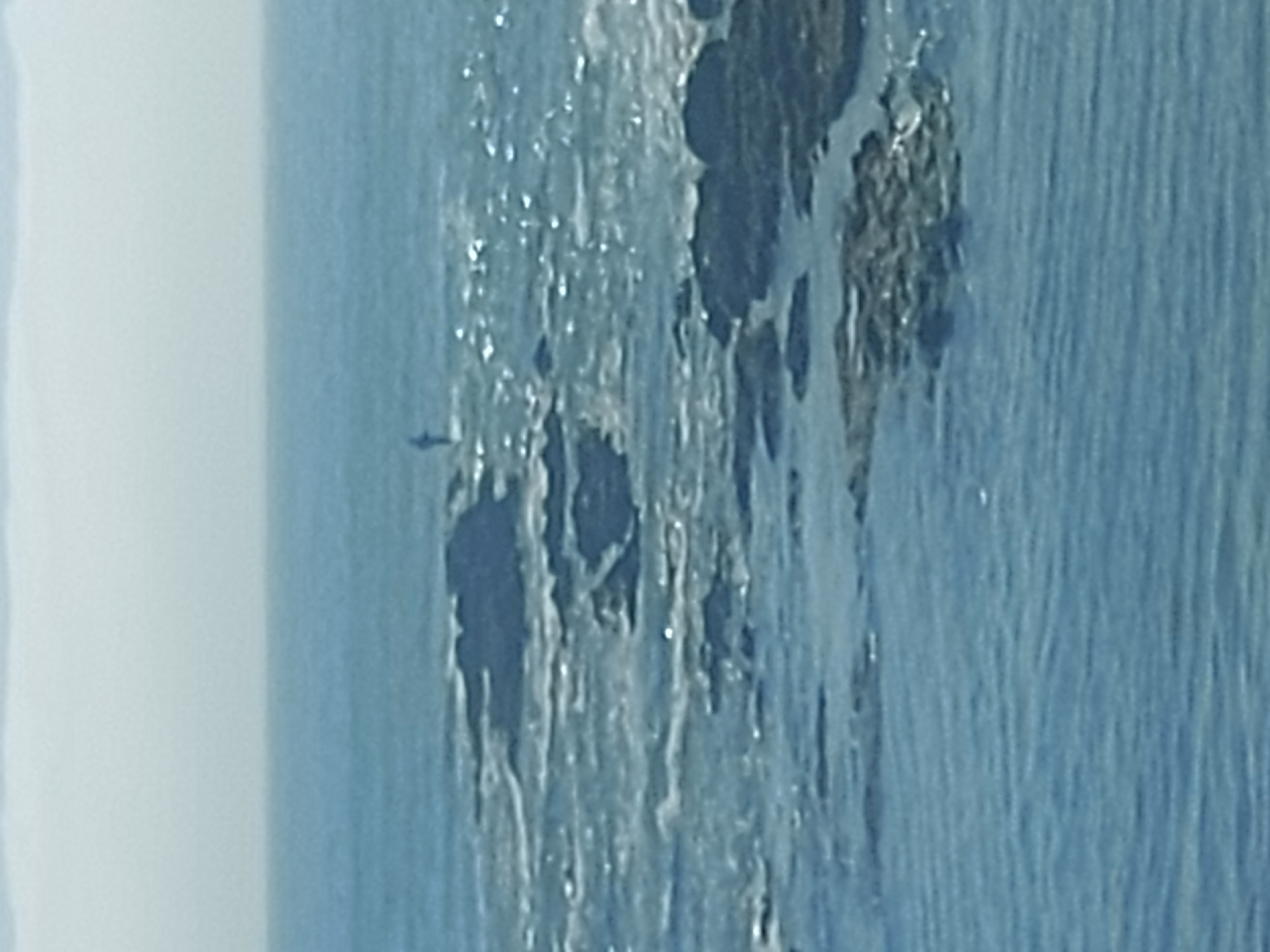 The image captures a coastal scene in Figueira da Foz, Portugal. Visible is a rocky outcrop or small island in the sea. Water is prominent, exhibiting textures and movement, likely waves. In the distance, a small aircraft or bird is barely visible. The environment appears maritime, with varying shades of blue.